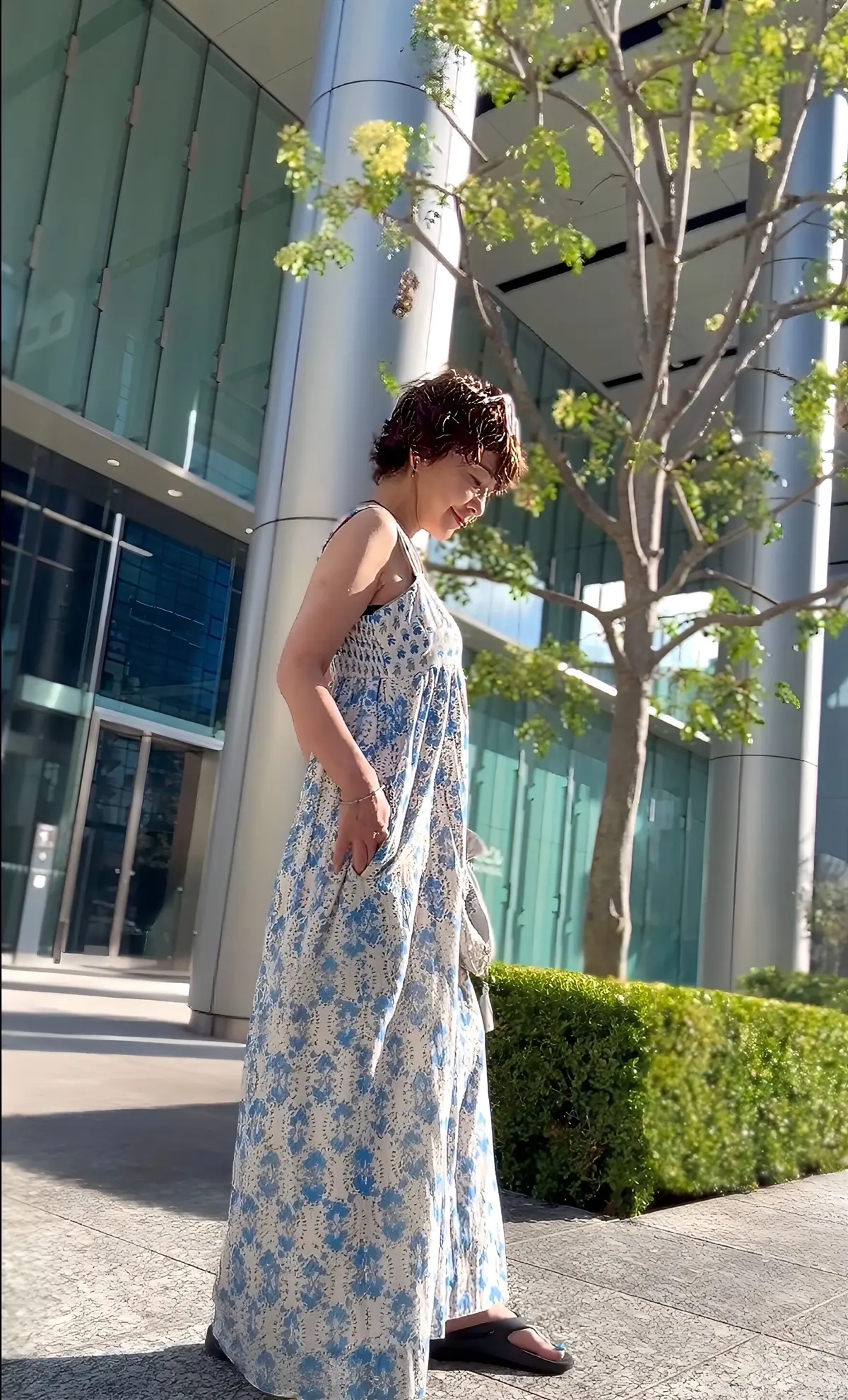 白い花柄のロングワンピースを着た女性が、近代的なビルの前でこちらを見ている。茶色のサンダルを履き、短い茶色の髪型。背景には緑の木々とガラス張りの建物がある。