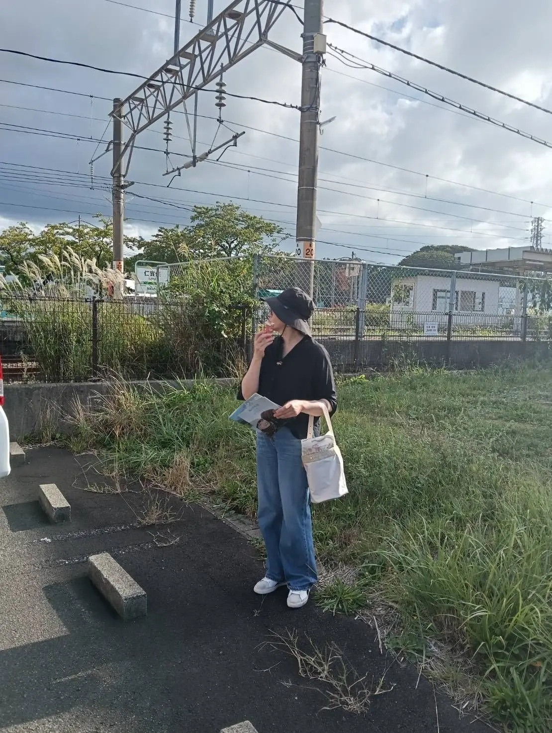 黒い帽子を被った女性が、鉄道の架線のある脇に立って本を読んでいる。青空を背景に、緑の草地とフェンスが見える。
