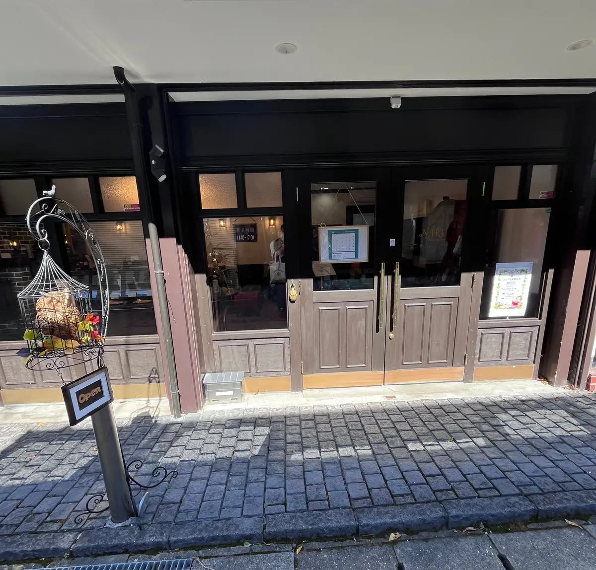 灰色の木製フレームの入り口を持つお店。鳥かご型の「Open」の看板が設置され、ガラス越しに店内の様子が見える。背景には茶色の建物がある。