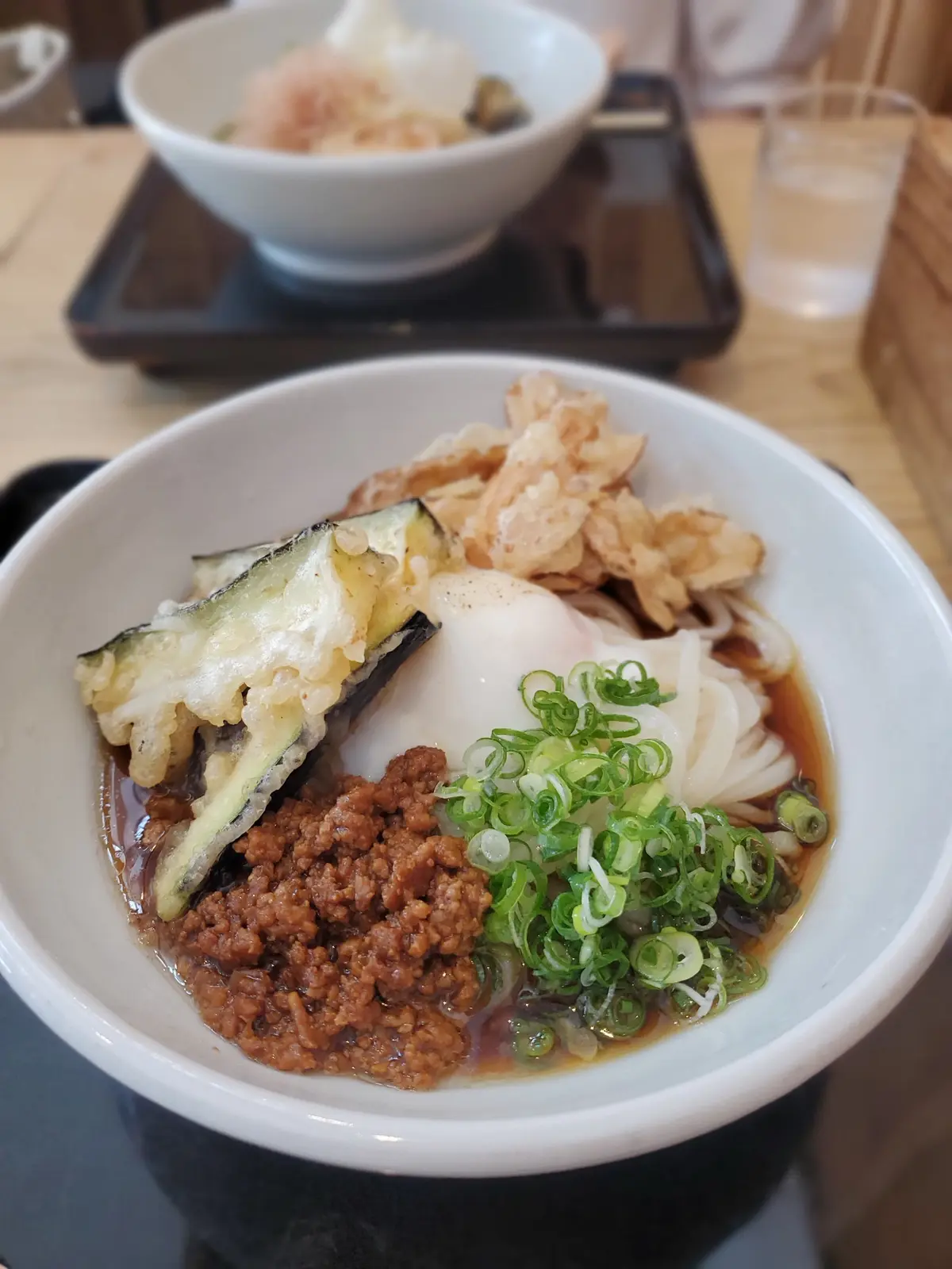 白い器に入った、温かい汁物。うどんのような麺に、ネギ、豚肉のそぼろ、野菜の天ぷらが乗っている。背景にはご飯と漬物が置かれた小鉢。