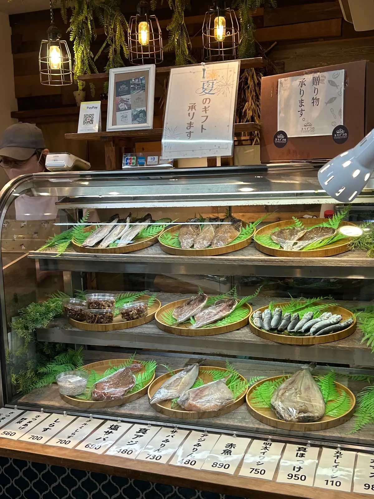 鎌倉レンバイ。木製のカウンターに並んだ、様々な種類の魚介類が展示されている様子。ガラスケース内に、生の魚や調理済みのものが盛り付けられ、緑の葉で飾られている。奥にはメニューや装飾品が置かれ、照明器具が吊り下げられている。