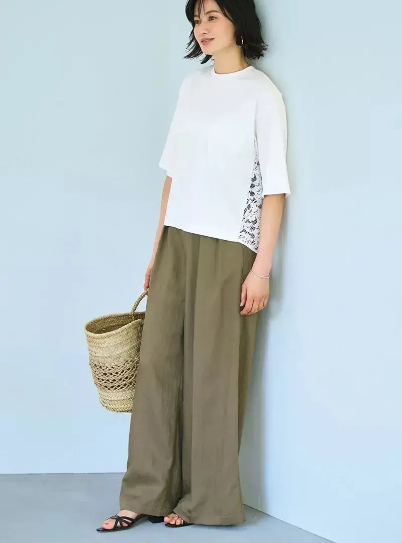 青い背景をバックに、女性が立ち上がって正面を見ている。白と柄物のトップスに、緑色のワイドパンツを着用。右手に編み込みのバスケットを持ち、足元には黒いサンダル。シンプルなスタイリングで、落ち着いた雰囲気が伝わる。