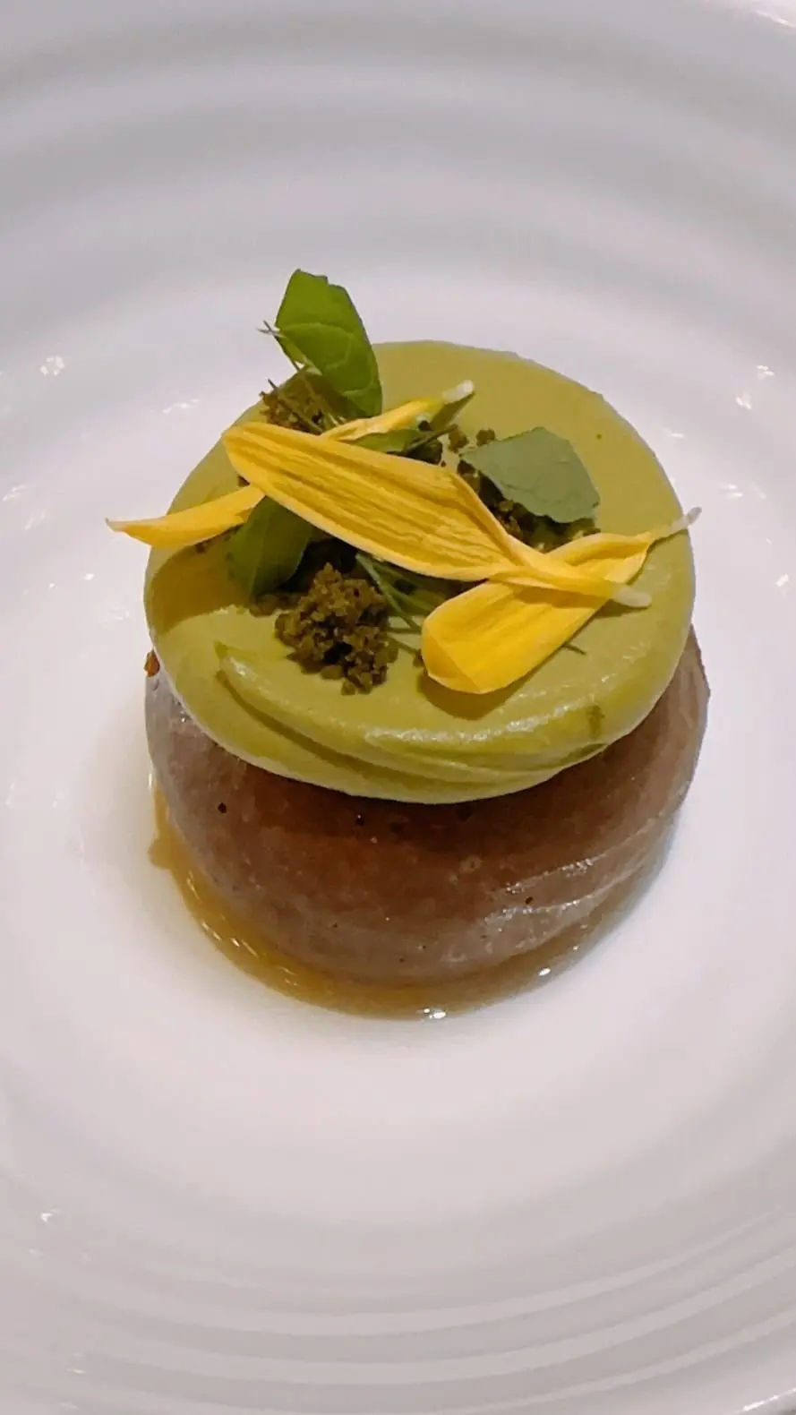 緑のクリームをのせた茶色いベースのデザートに、黄色い花と緑の葉が飾られている