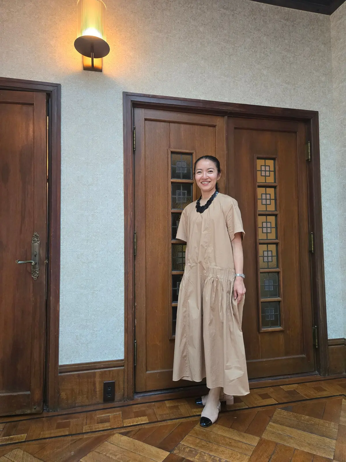 ベージュのワンピースと黒のアクセサリーを身につけた女性が、木製のドアの前で微笑んでいます。壁には壁灯が取り付けられています。