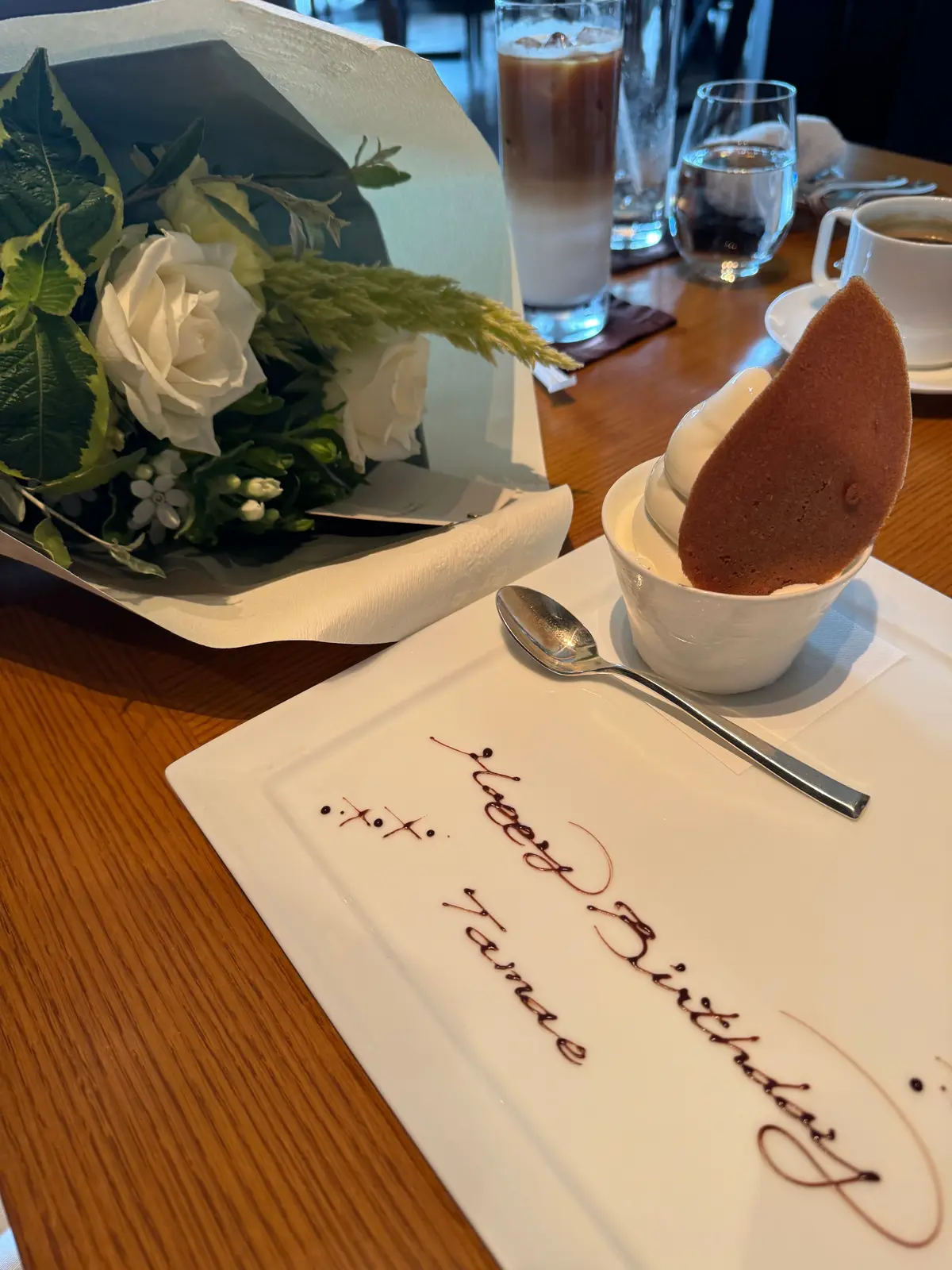 木製のテーブルに置かれた白いお皿に、手書きで「Happy Birthday Tamae」と書かれたメッセージと、白い花束、ムース、コーヒー、飲み物が写っている。
