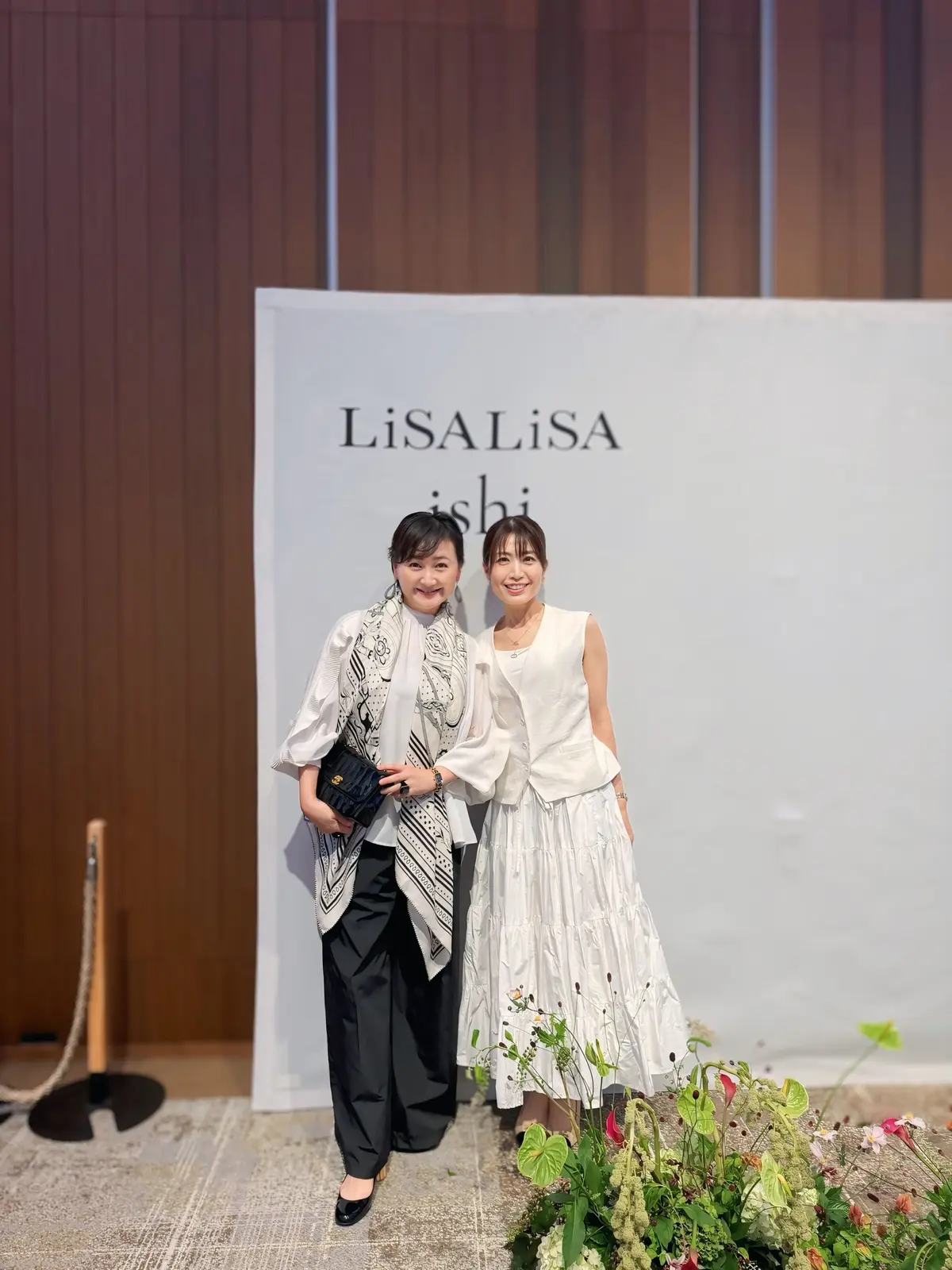 LiSA LiSAのロゴを背景に、2人の女性が並んで立っている写真。左の女性は黒いパンツスーツに柄物のスカーフ、右の女性は白いフレアスカートのトップスを着用。手前に花束が置かれている。