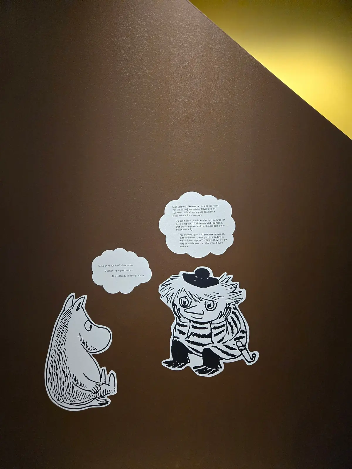 Moomin（ムーミン）のキャラクター、ムーミンパパとムーミントロールのイラストが壁に貼られている。白地に黒の線で描かれ、それぞれに短いテキストが添えられている。背景はベージュ色の壁。