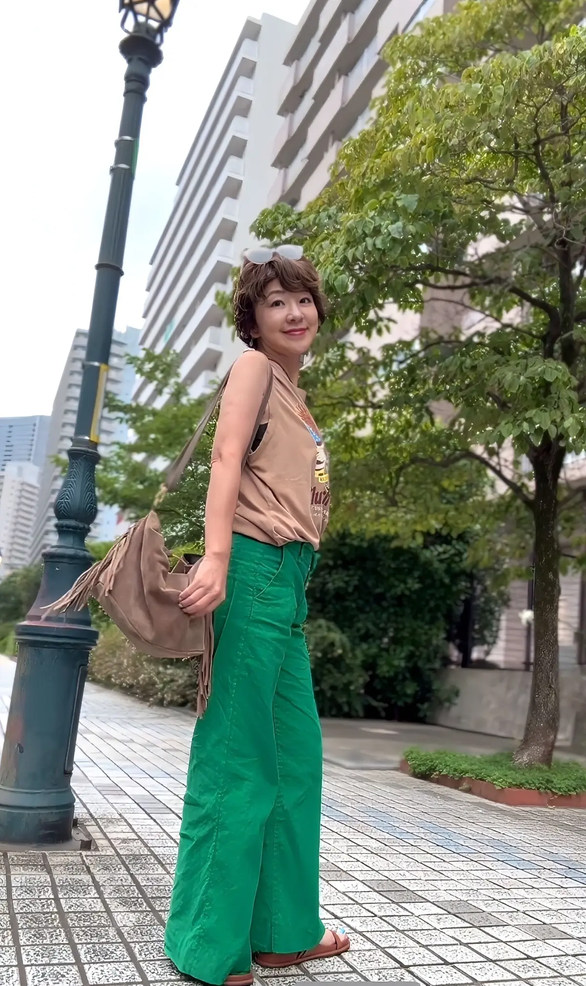 緑色のワイドパンツに茶色のタンクトップを着た女性。茶色のリュックを背負い、笑顔でこちらを見ている。背景には高層ビルと緑豊かな木々が見える。