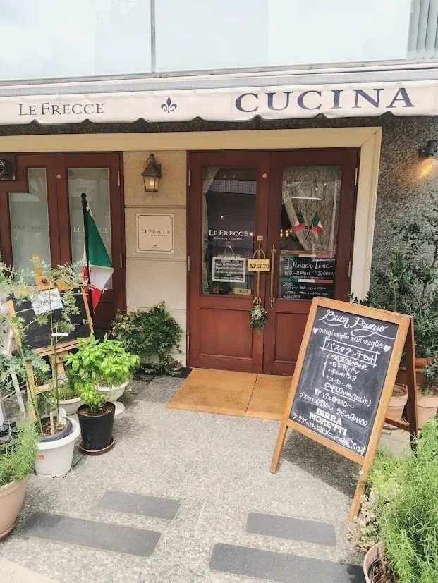 木製のドアと窓のあるイタリア料理店「Le Frecce Cucina」の入り口。イタリア国旗が飾られ、手書きのメニュー看板が置かれている。入り口付近には植栽が配置され、落ち着いた雰囲気を醸し出している。