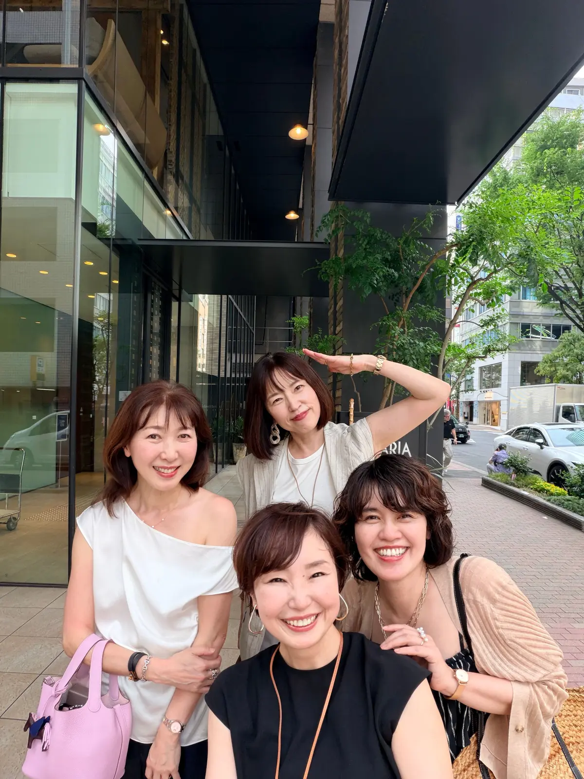 5人の女性が屋外で笑顔で写る集合写真。建物入口の前で、左から2人、3人が白いトップス、中央が黒いトップス、右端がベージュのカーディガンを着ている。右端の女性が腕を上げ、ポーズをとっている。