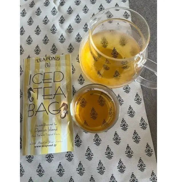 Teapond社の「パッションアイランド」アイスティーバッグと、茶色の模様が入ったテーブルクロスの上に置かれた2つのグラスに入った黄色いお茶。