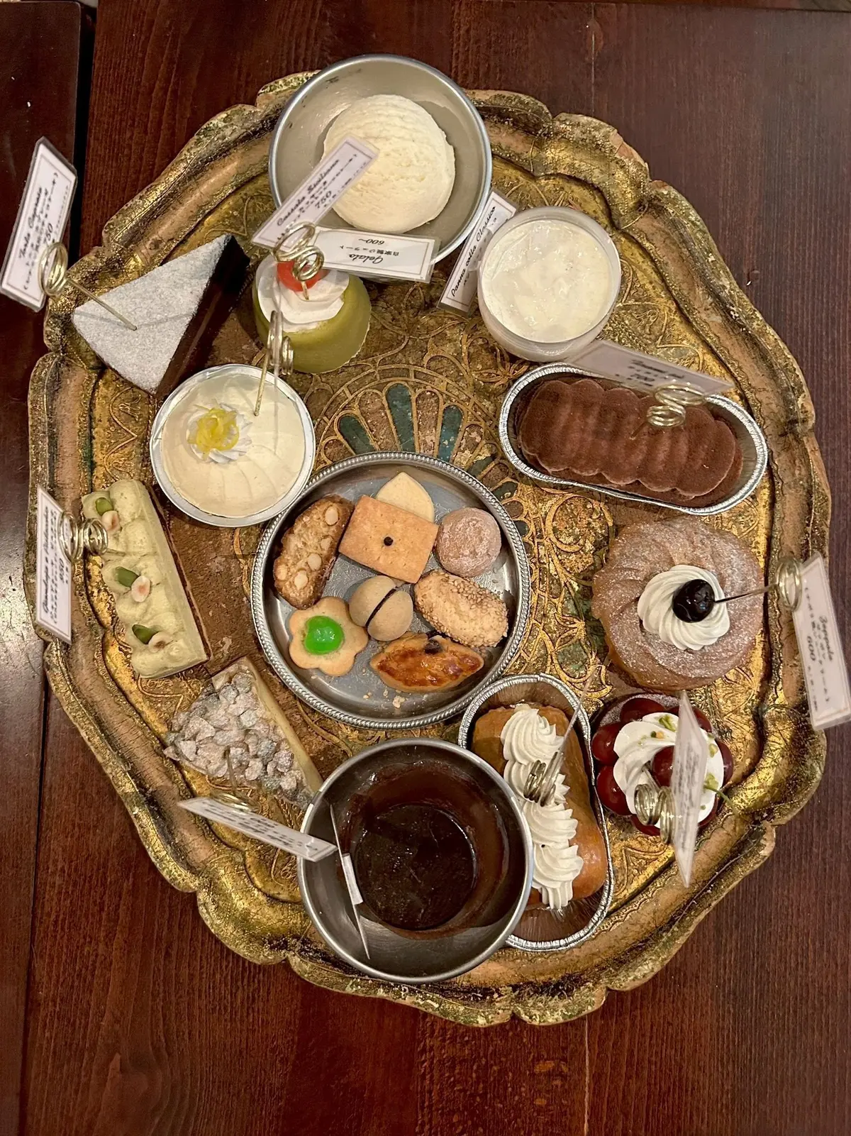 金色の装飾が施されたお盆に、様々な焼き菓子やクリーム、チョコレートなどが盛り付けられている。中央にはクッキーの盛り合わせ、周囲には小分けにされたデザートが並び、上品なティータイムの様子を伝えている。