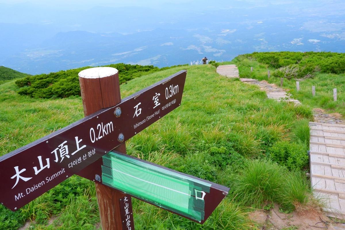 山頂の緑豊かな草原に設置された木製の案内板で、左方向に「大仙山頂 Mt. Daisen Summit 0.2km」、右方向に「石屋屋敷 Ishiya Shiki 0.3km」と表示されている