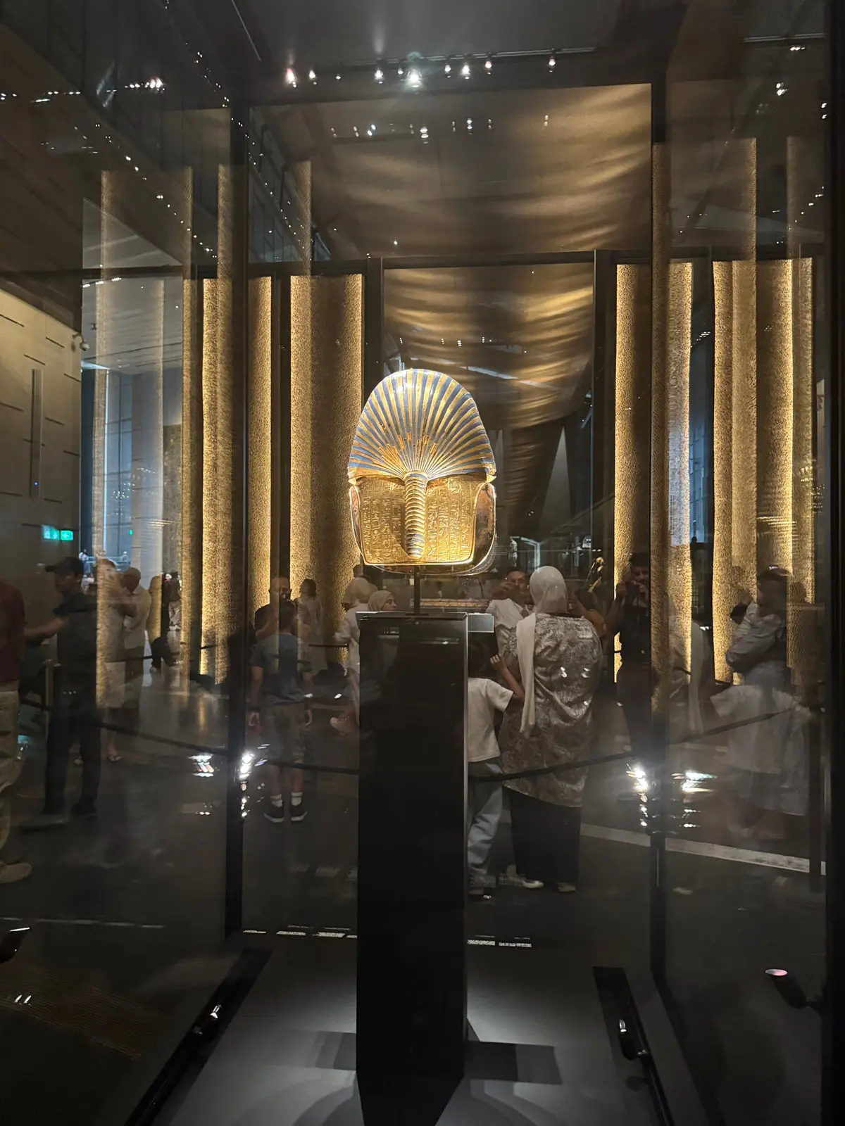 エジプトのツタンカーメン王の黄金のマスクが展示された空間。ガラスケース内に展示され、周囲には金色の柱が立ち並び、多くの人が見学している。