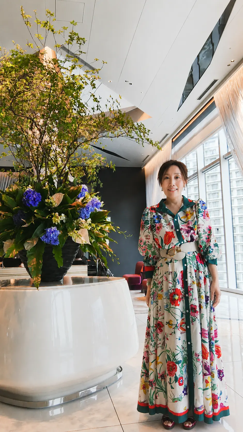 女性が花模様のロングドレスを着て、モダンなロビーの窓際で立っている。左側に青い花が咲く大きな花瓶がある。