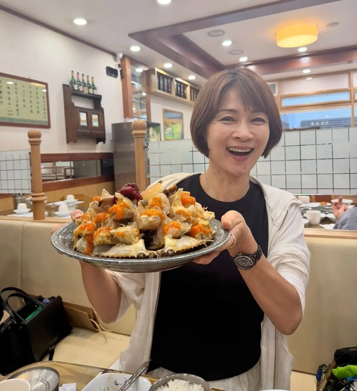 レストランのテーブルで、驚いた表情の女性が、たくさんの蟹料理が盛られた皿を持っている。皿はグレーの金属製で、蟹の身や卵、赤い唐辛子が散らばっている。背景にはテーブル席と窓が見える。