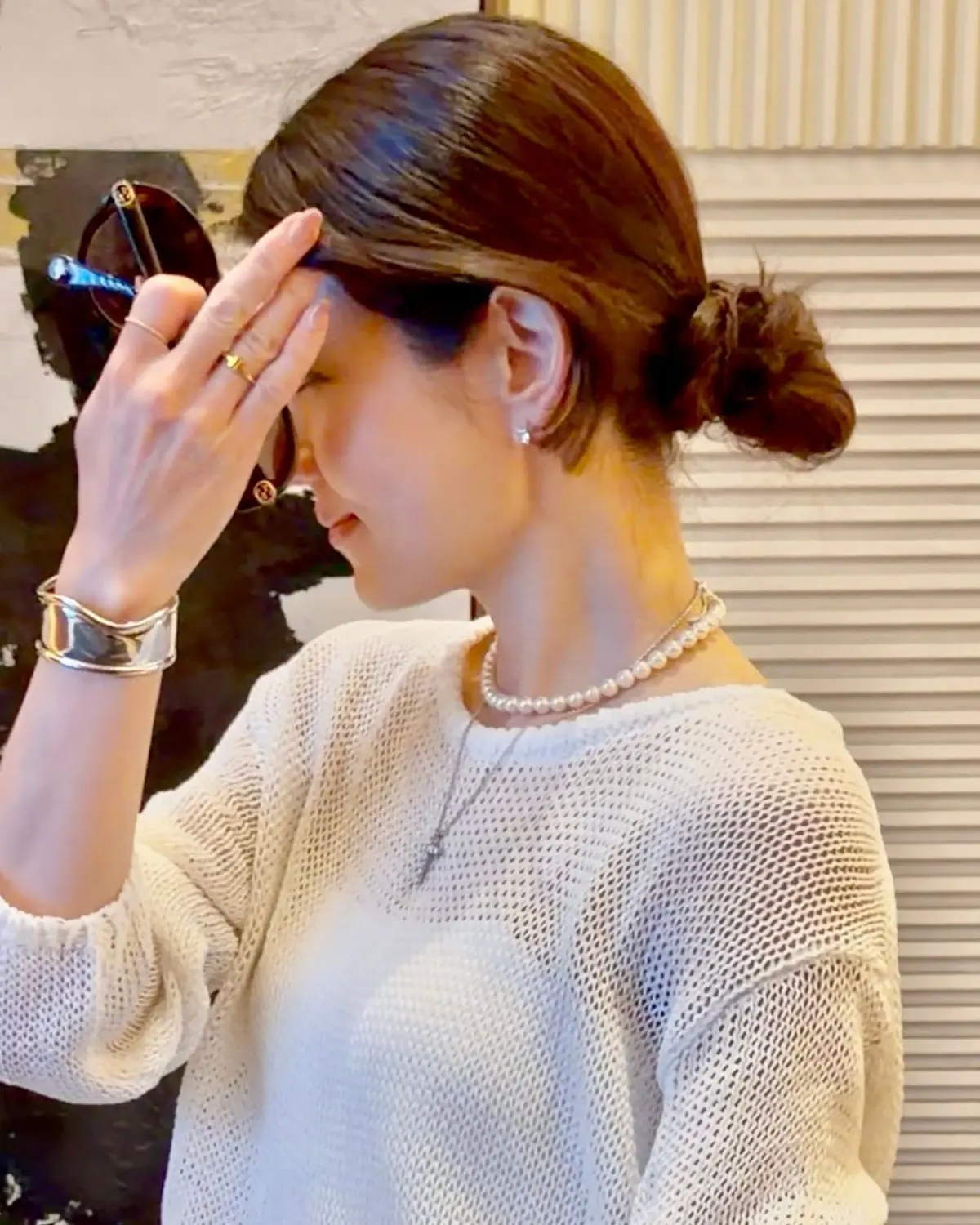 女性が白い編み物セーターを着て、真珠のネックレスと銀のブレスレットを身につけ、片方の手でサングラスを調整している。背景には抽象的な絵画がかけられている。
