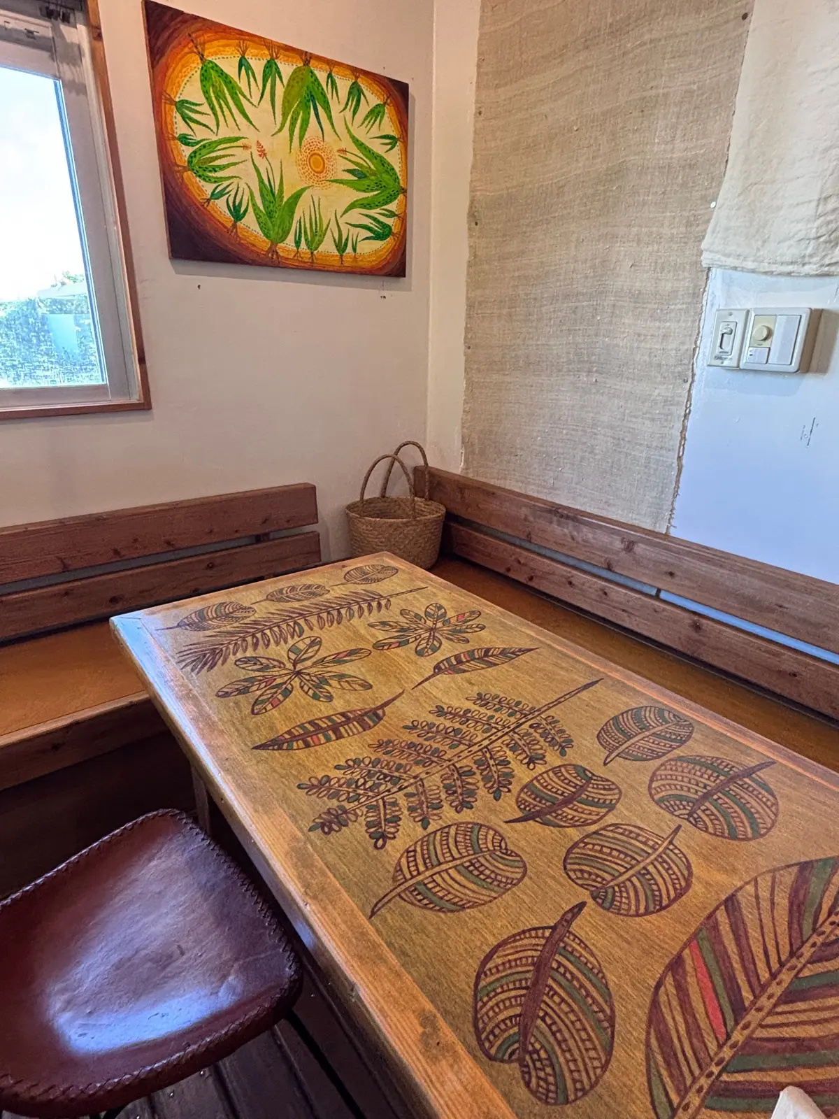 木製のテーブルに、葉や植物をモチーフにした模様が描かれている。壁には植物画が飾られ、窓からは光が差し込んでいる。右側には麻のカーテンと電気のスイッチがある。