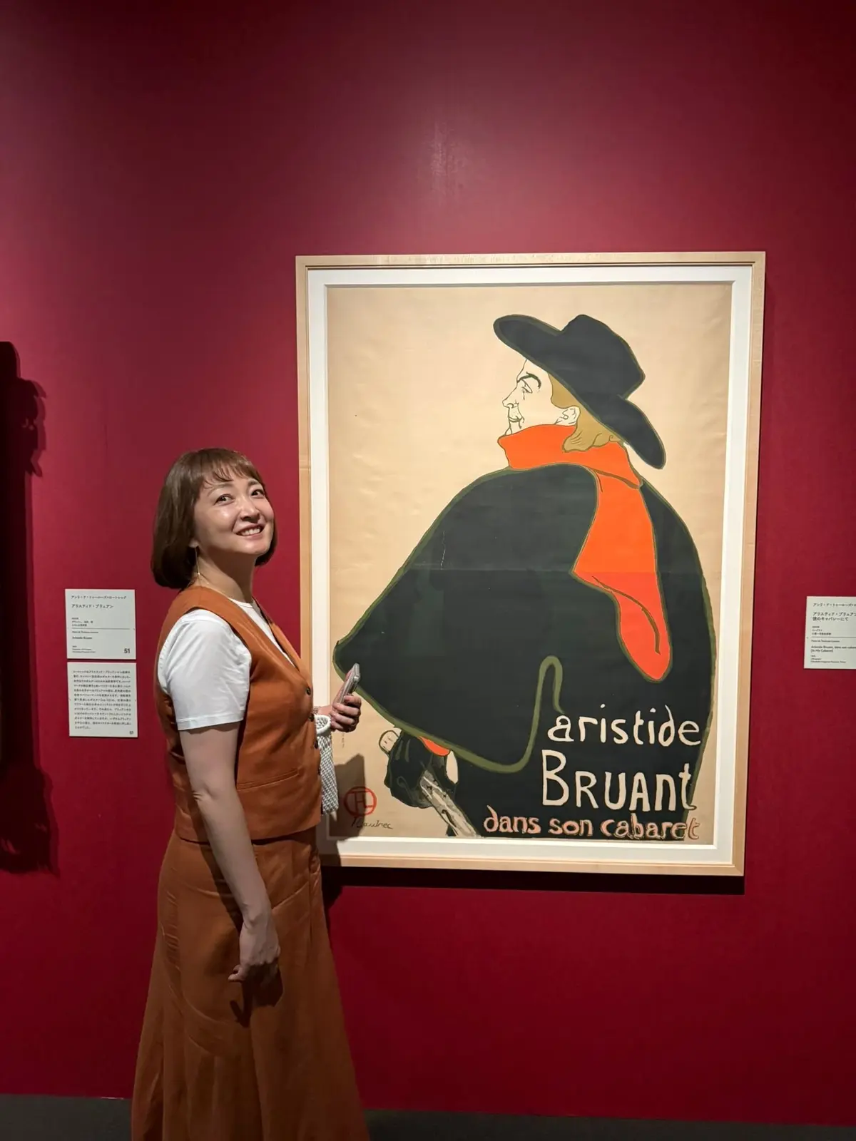 赤い壁の前で、女性がアート作品を背景に微笑んでいる。作品は、帽子を被った人物のポスターで、黒、オレンジ、ベージュの配色。女性はオレンジ色のベストとスカートを着用。