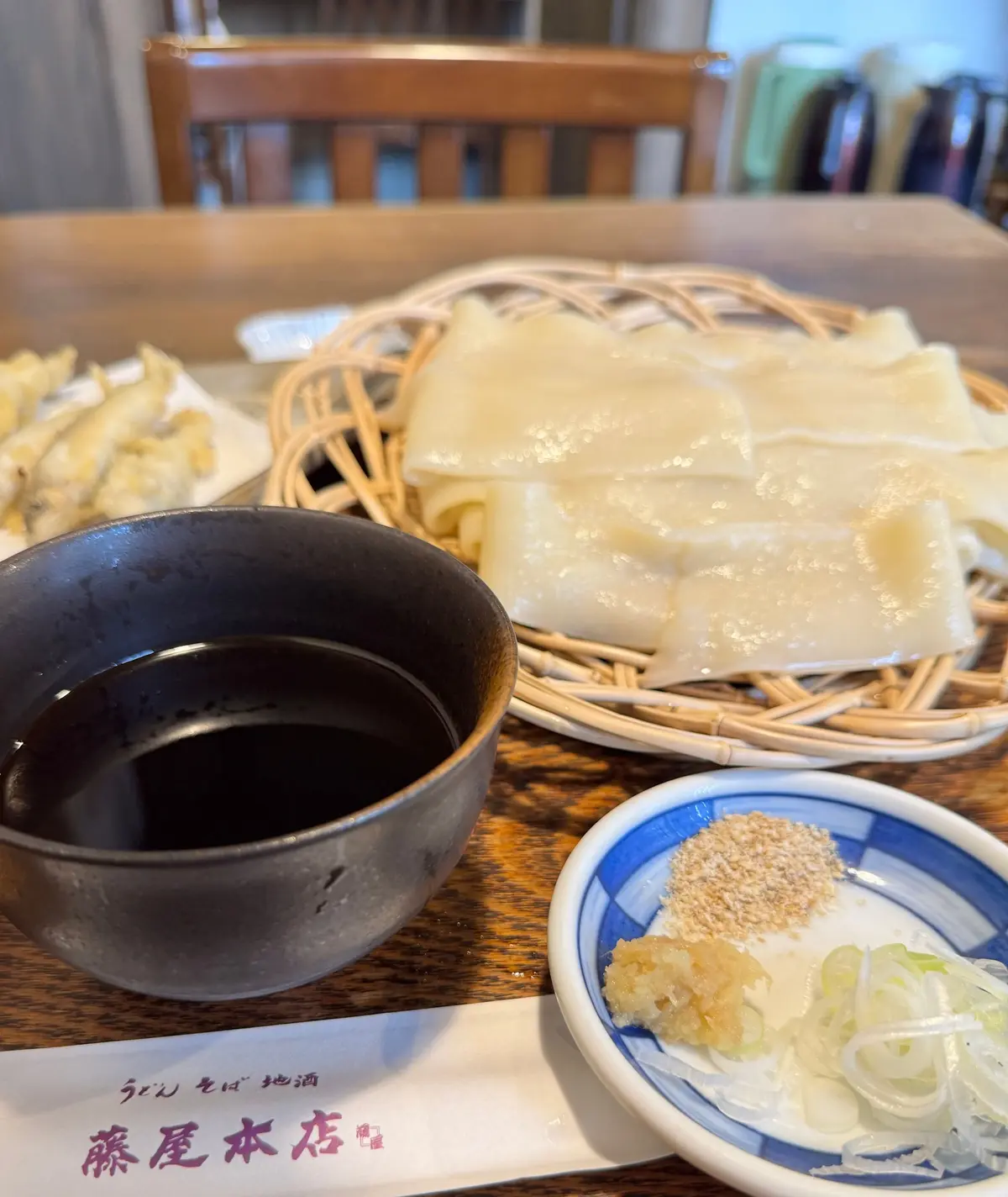 木製のテーブルに置かれた、中華料理の点心と付け合わせの盛り合わせ。点心は白く、蒸されており、竹製のザルに盛られている。左には黒い醤油、右には白髪ねぎと生姜の和え物が青い皿に入っている。