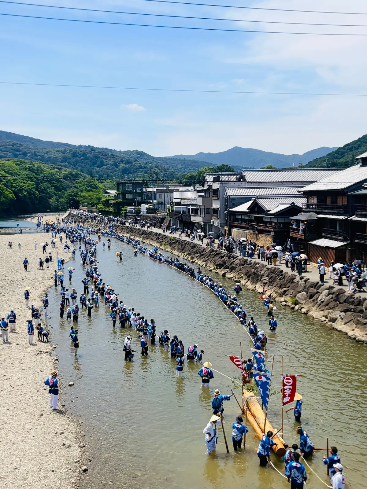 浅瀬に沿って、青い衣装をまとめた人々が長い列を作って歩いている様子。背景には伝統的な家並みと山々が広がり、晴天の日の風景。