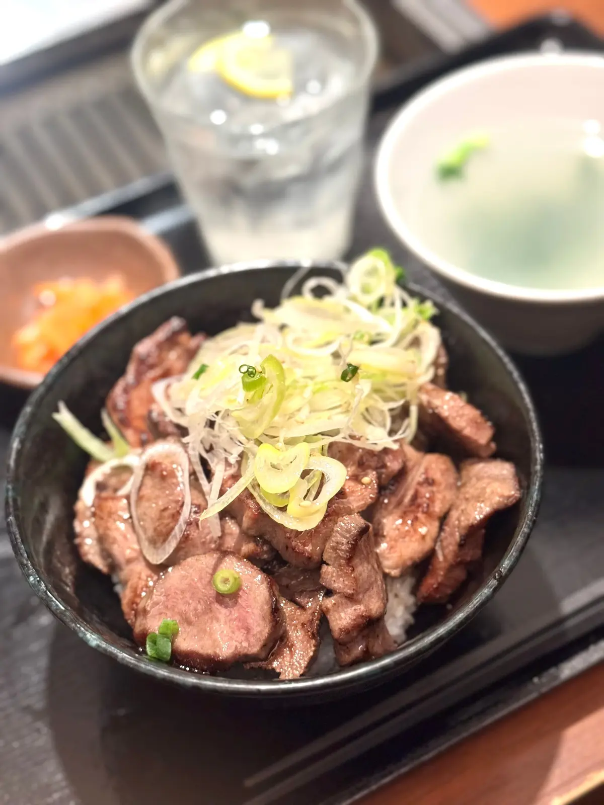 黒い鉢に牛肉のステーキ、細切りのネギと白菜が乗せられ、横に水とスープの小鉢が並ぶ