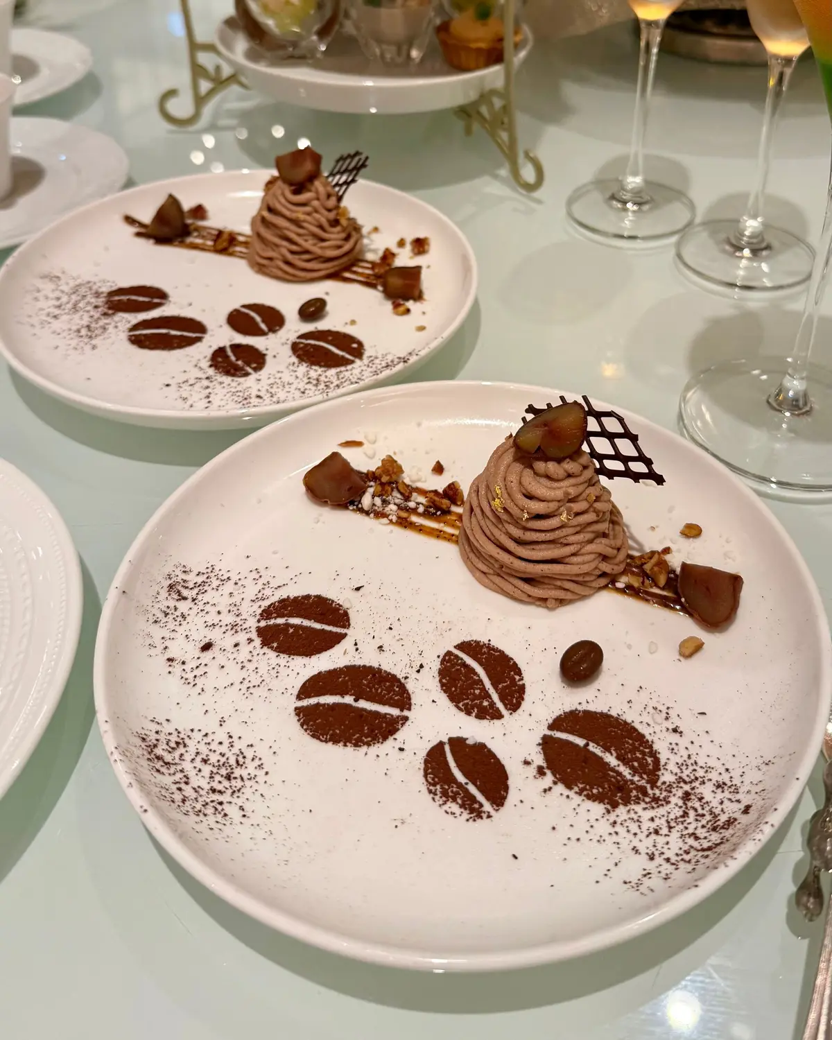 白磁の皿に盛り付けられた、コーヒー風味のデザート。中央には茶色のムース、周りには砕いたナッツやチョコレート、金粉が散りばめられている。皿にはコーヒー豆の形にココアパウダーが描かれ、上品な印象。背景にはグラスとケーキスタンドが見える。