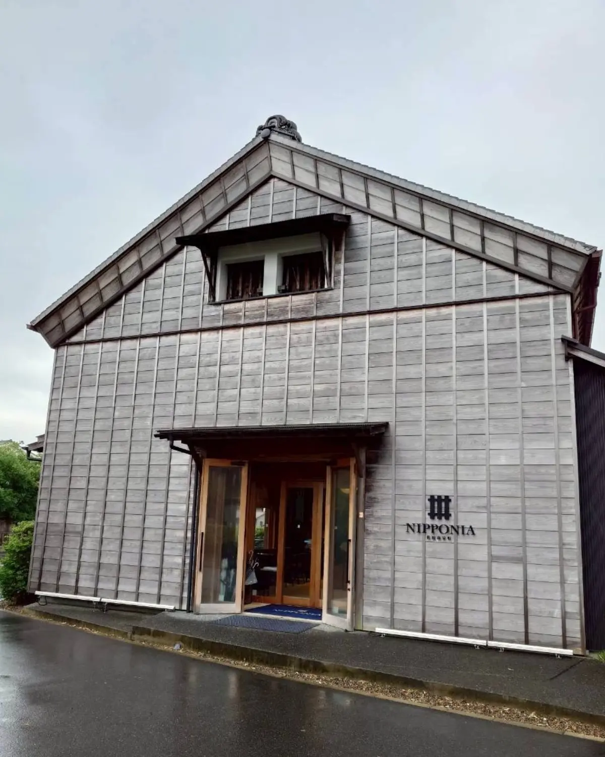 木造の建物は灰色で、三角屋根と木製の外壁を持つ。中央にガラスのドアがあり、右側に「佐原商家町ホテルNIPPONIA」の文字が記されている。