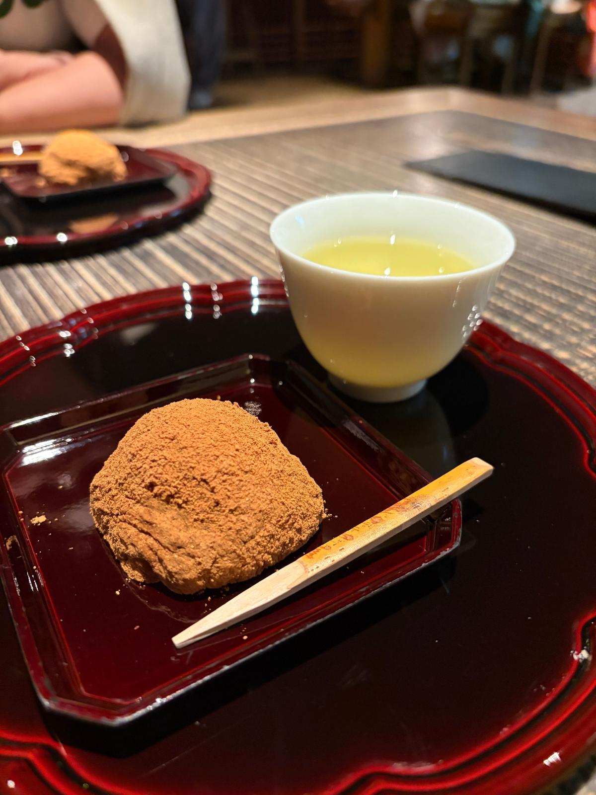 赤いプレートに茶色い粉状のスイーツと白いカップに入った黄色い液体があり、木製のナイフが添えられている
