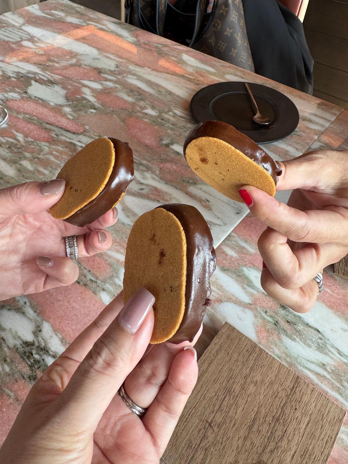 3つのチョコレートコーティングされたビスケットが、人々の手によって運ばれている。それぞれが半分に割れており、内部のスポンジのような生地が見えている。背景には、マーブル模様のテーブルと、ルイ・ヴィトンのバッグが見える。
