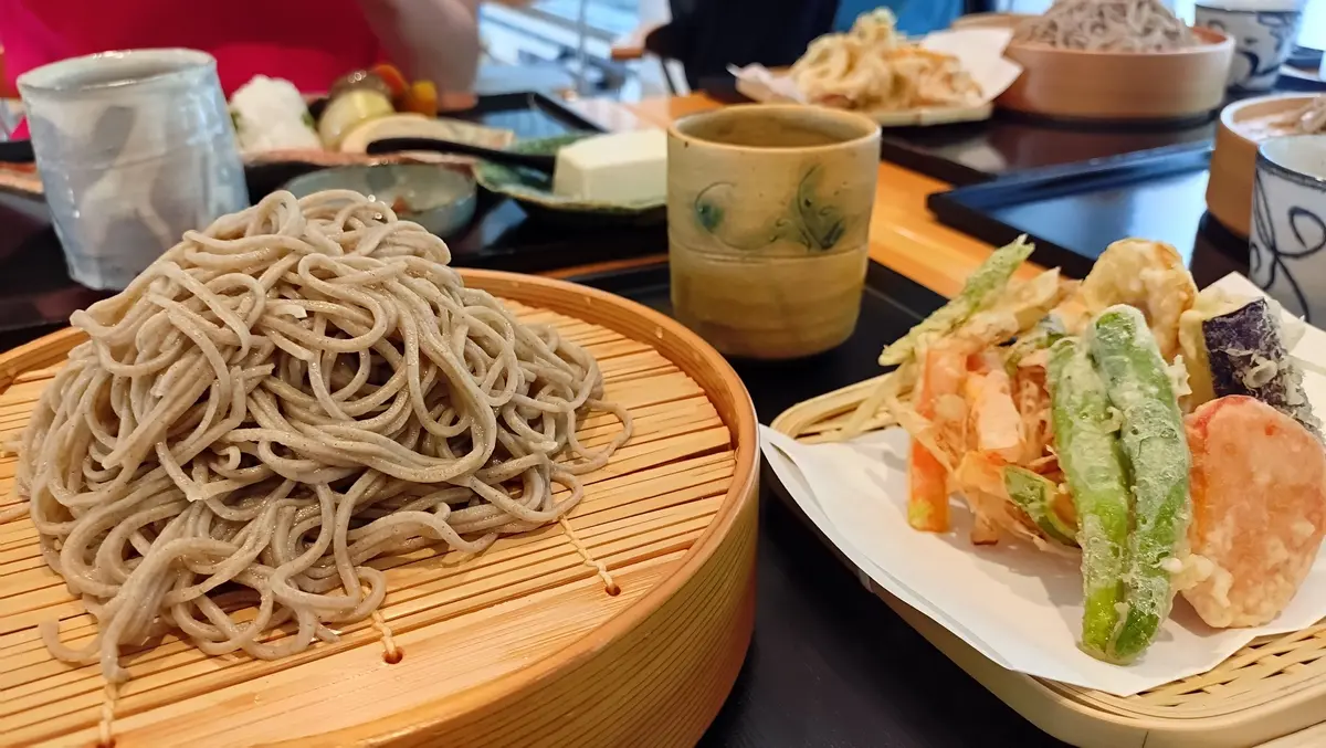 竹製の盛り付け皿に盛られた蕎麦と、天ぷらの盛り合わせ。蕎麦はねじれた形状で、天ぷらは野菜や魚介がカラッと揚げられている。背景には茶碗やグラス、その他の料理が見える。