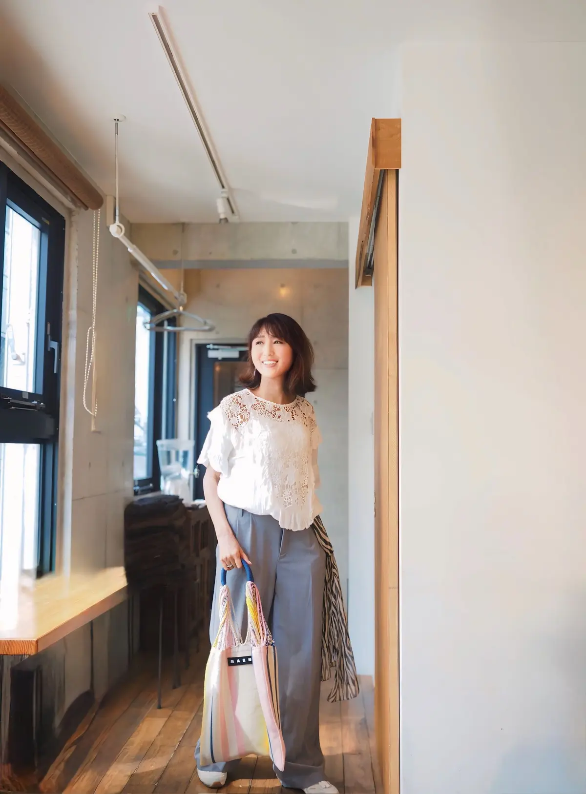 白いレースのトップスにグレーのワイドパンツを着用した女性が、カラフルなトートバッグを持って立っている。背景には窓と木製の床が見える、明るい室内。