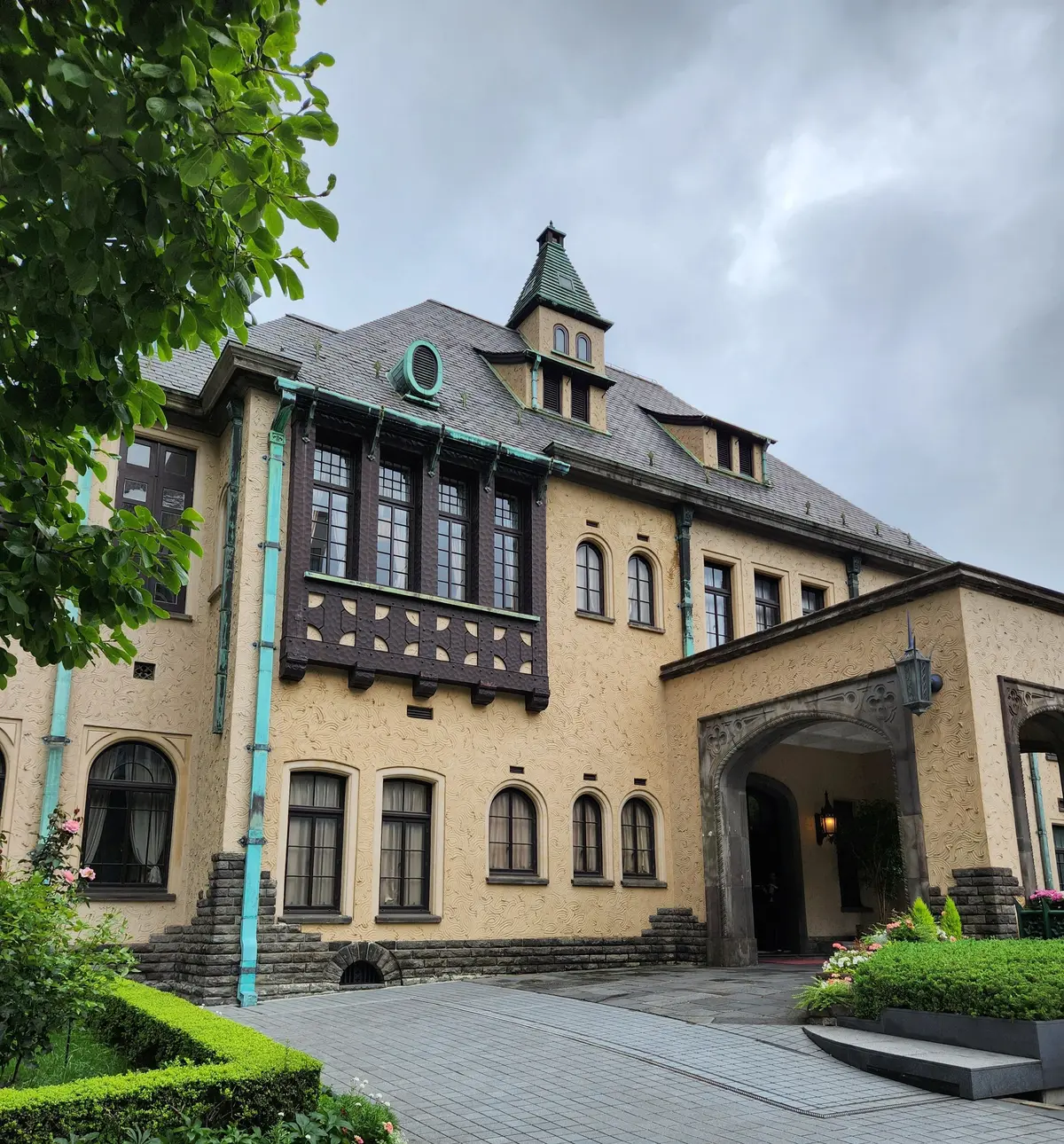 ベージュの外壁と緑の装飾が施された2階建ての洋館。石造りの階段と花壇が広がる石畳の前に建つ。窓にはカーテンがかけられ、左右に緑の木々が映える。