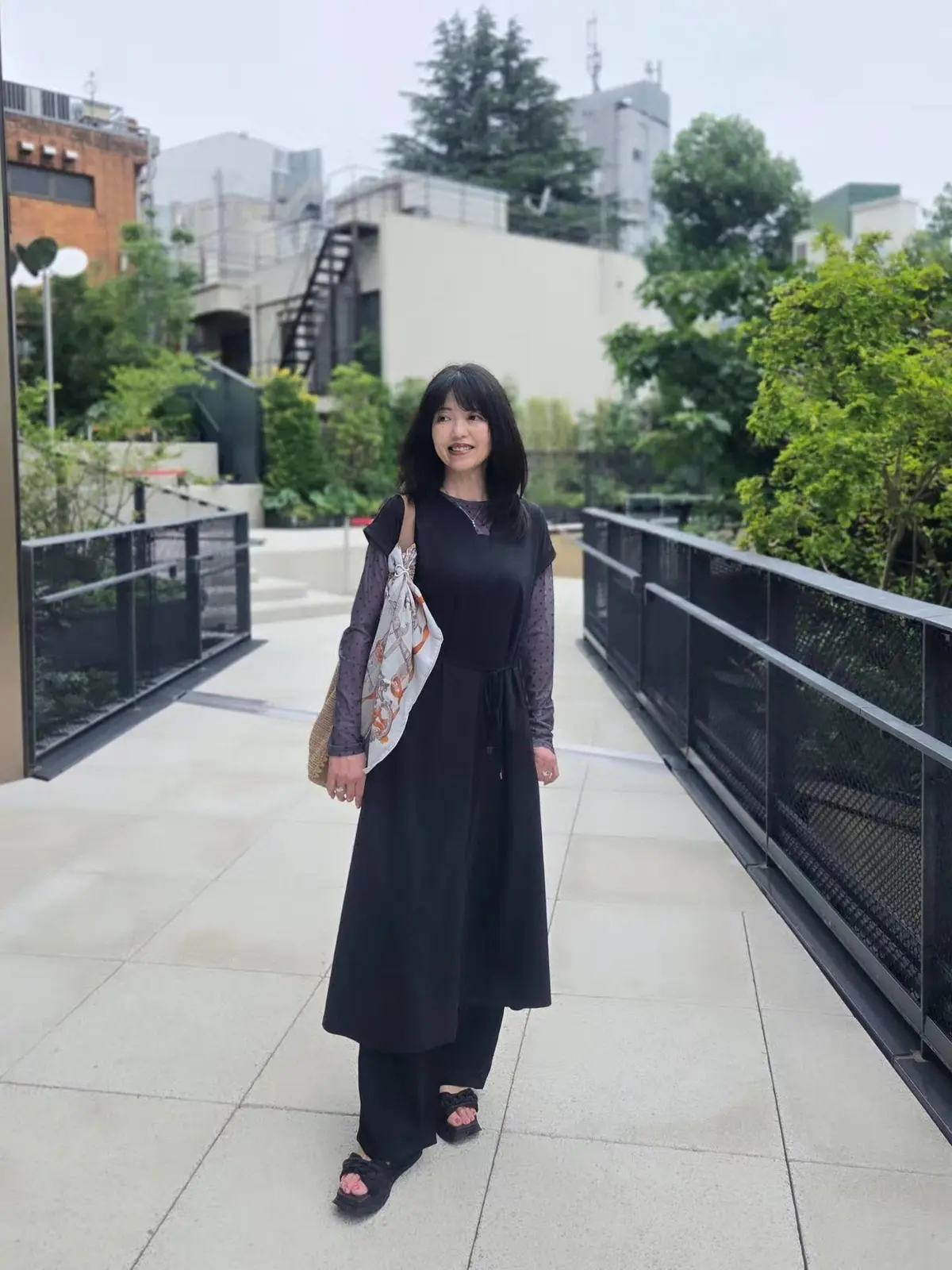 黒のロングドレスを着た女性が屋外の通路を歩く。左手に花模様のスカーフがかけられたカゴバッグを所持。背景には緑の木々や建物が並ぶ。