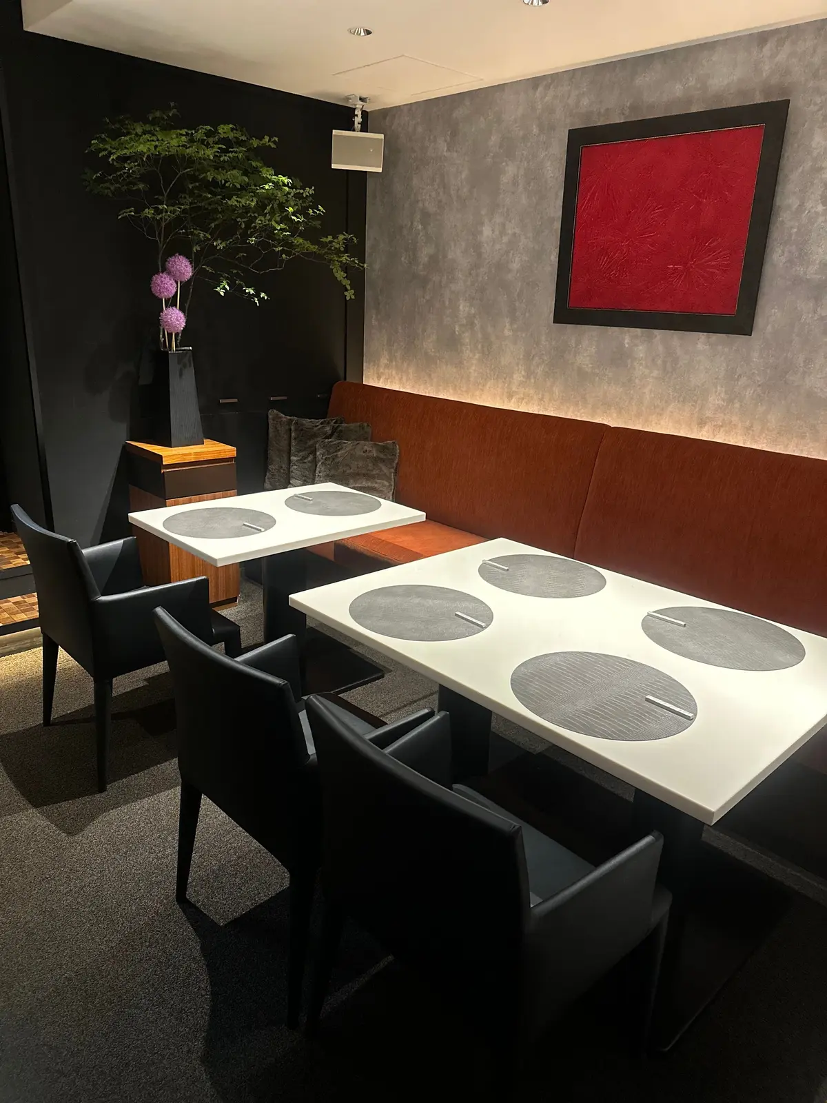 白い円形のテーブルに黒い椅子が配置され、木製のベンチソファが隣接するモダンなダイニングスペース。壁には赤い絵画と緑の観葉植物が飾られ、木製の小テーブルに紫の花が置かれている。