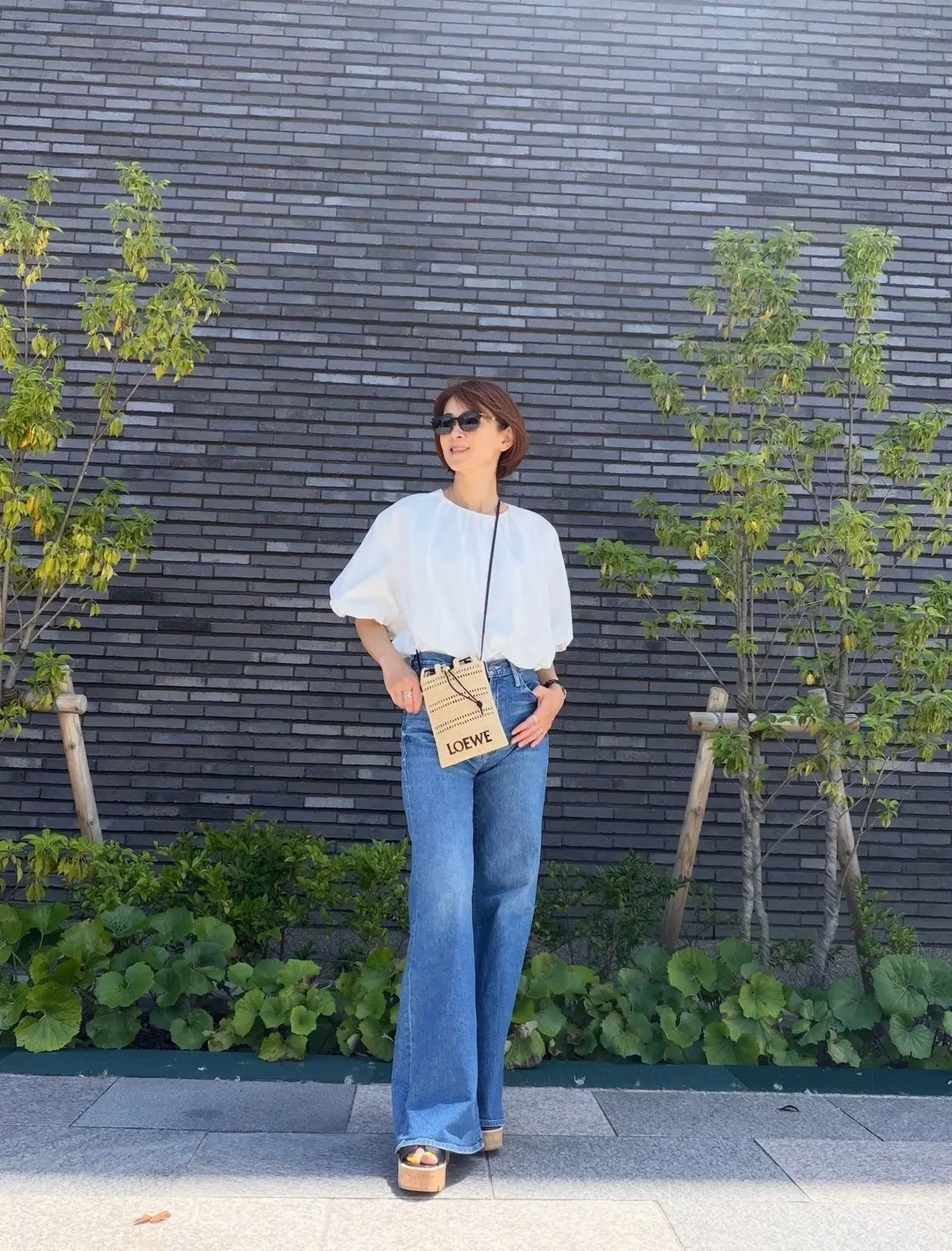 白のふんわりした袖のブラウスとデニムパンツを着た女性が、壁の前で立ち、LOEWEのショルダーバッグを下げている