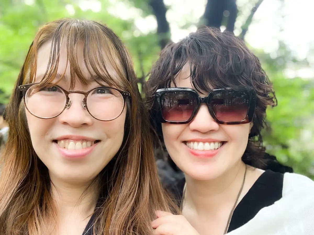 長髪と短髪の二人の女性が公園で並んで笑っている。左の女性は丸いメガネをかけ、右の女性はサングラスをかけ、背景には木々がぼんやりと見える。