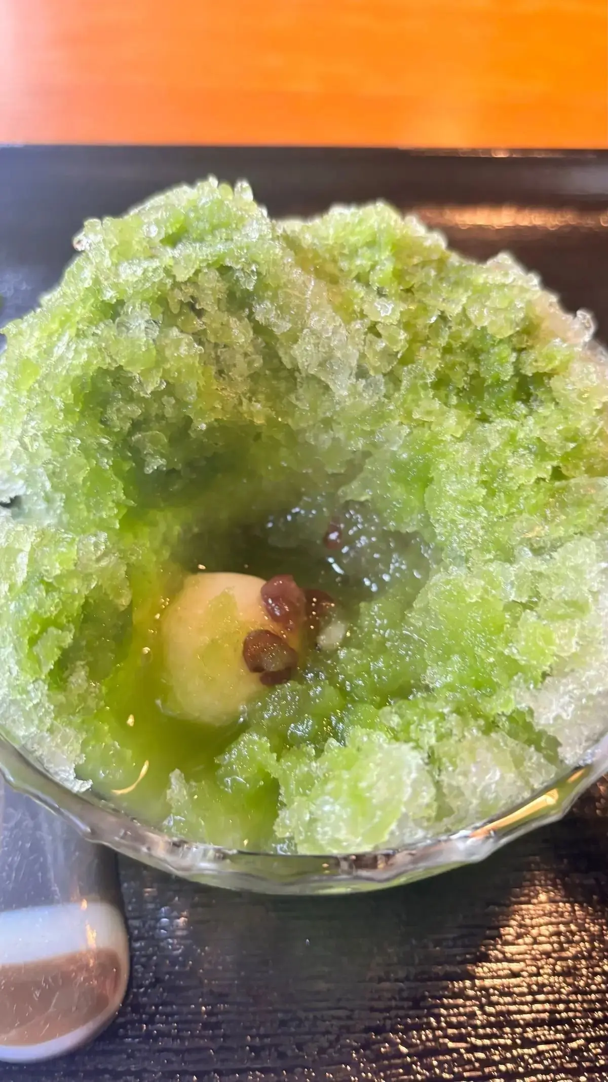 透明な器に入った、緑色のかき氷。中央には、梨と赤褐色の豆が入っている。表面は粗く、砂糖のような結晶で覆われている。背景はオレンジ色。