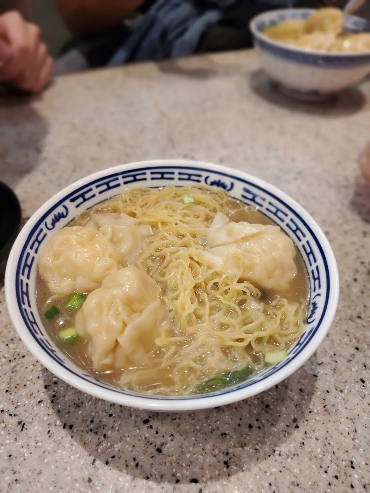 白い青い模様の器に、細麺と数個の肉まんが入ったスープ。緑の野菜が浮かんでいる。
