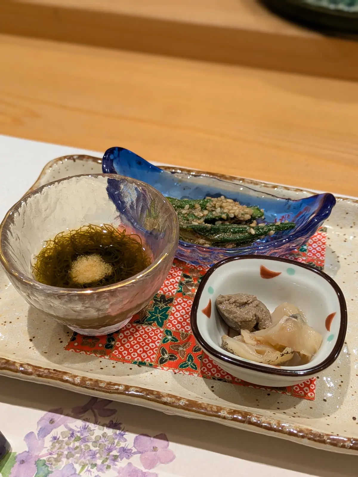 陶器の小鉢3つと長方形の容器が赤い花模様の敷物に並ぶ。左の鉢には海藻と卵黄、右の鉢には白身の魚と豆腐、中央の容器には枝豆とご飯が盛られている。