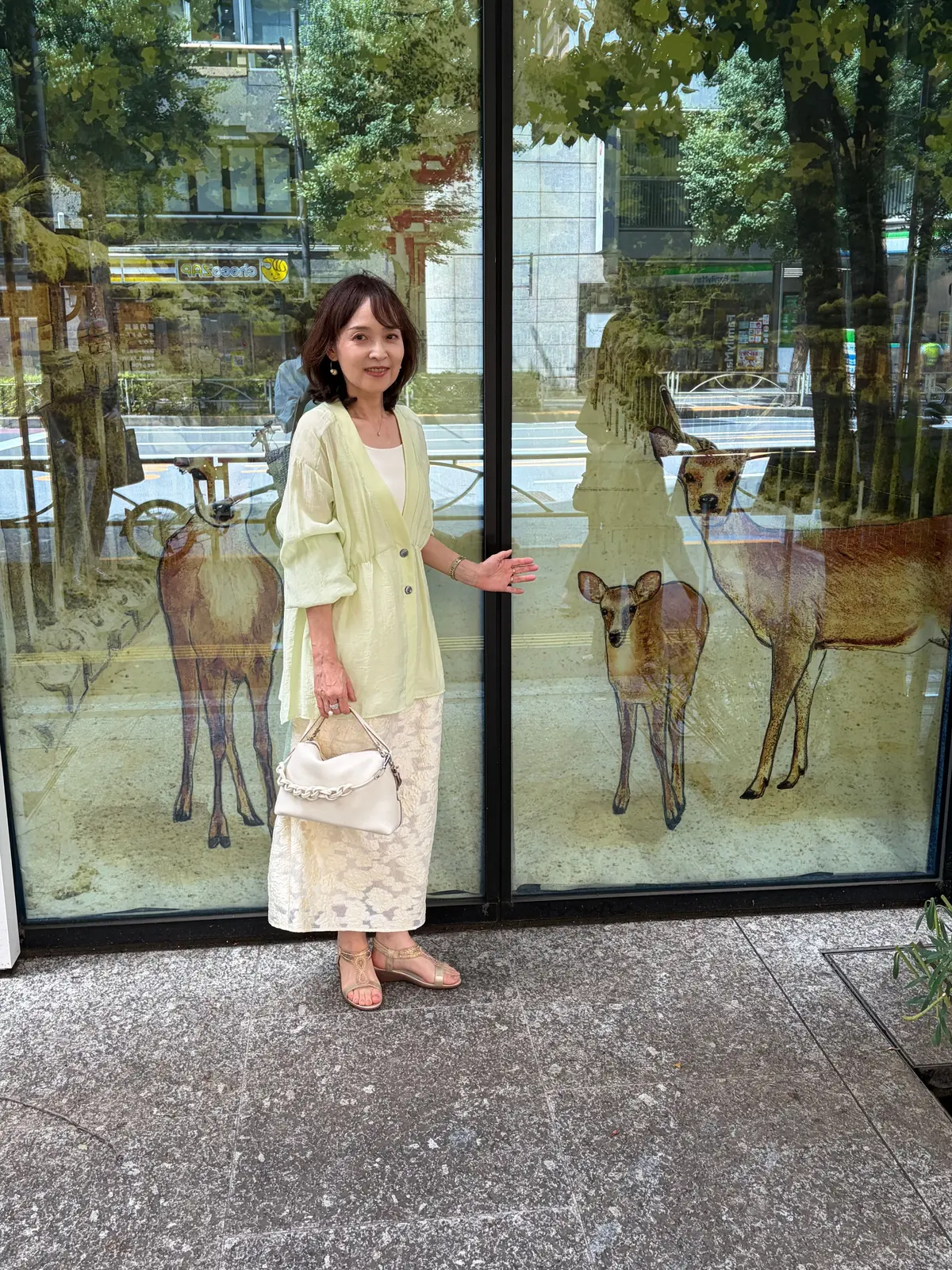 緑色のカーディガンに白いスカート、サンダルを身につけた女性が、鹿の絵が描かれたガラス窓の前に立っている。右手に白いハンドバッグを持つ。背景には建物と木々が見える。