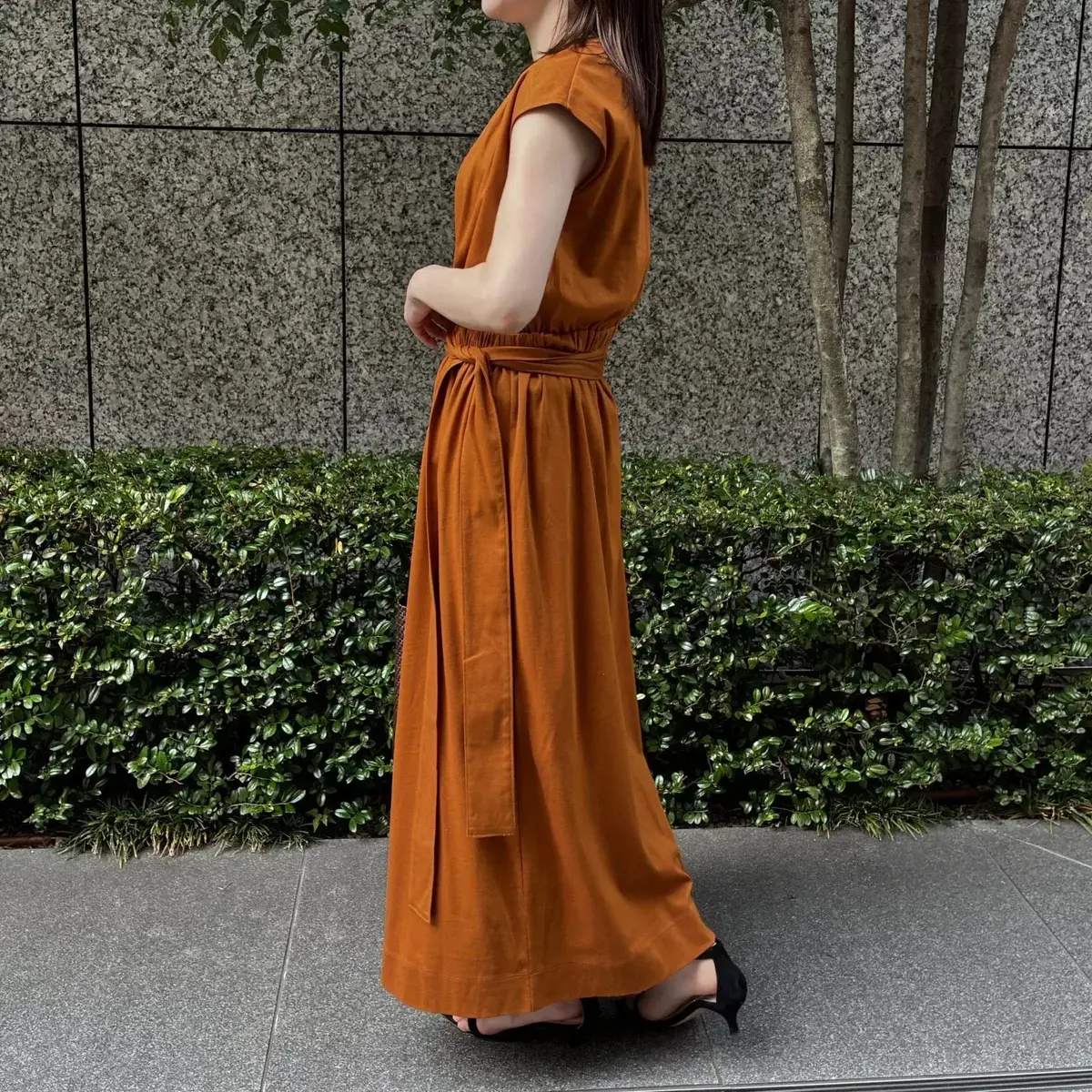 茶色のロングドレスを着た女性が、緑の植栽を背景に立っている。カフタンタイプのワンピースで、ウエストに紐がある。黒いパンプスを履いている。