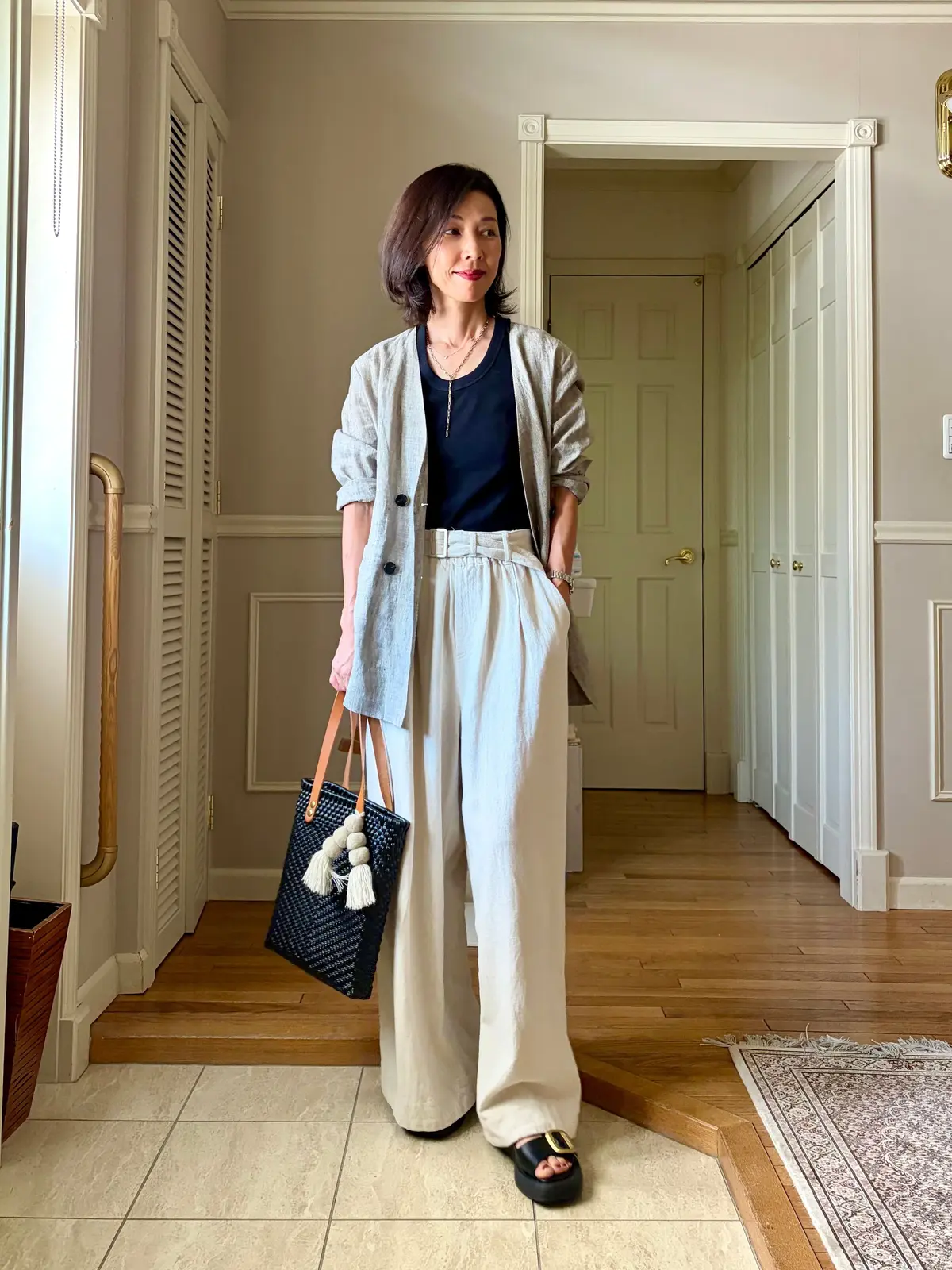 ベージュのジャケット、黒のTシャツ、同系色のワイドパンツを着用した女性が立っている。黒いサンダルを履き、編み込みのトートバッグを持つ。背景には白いドアと木製の床が見える。