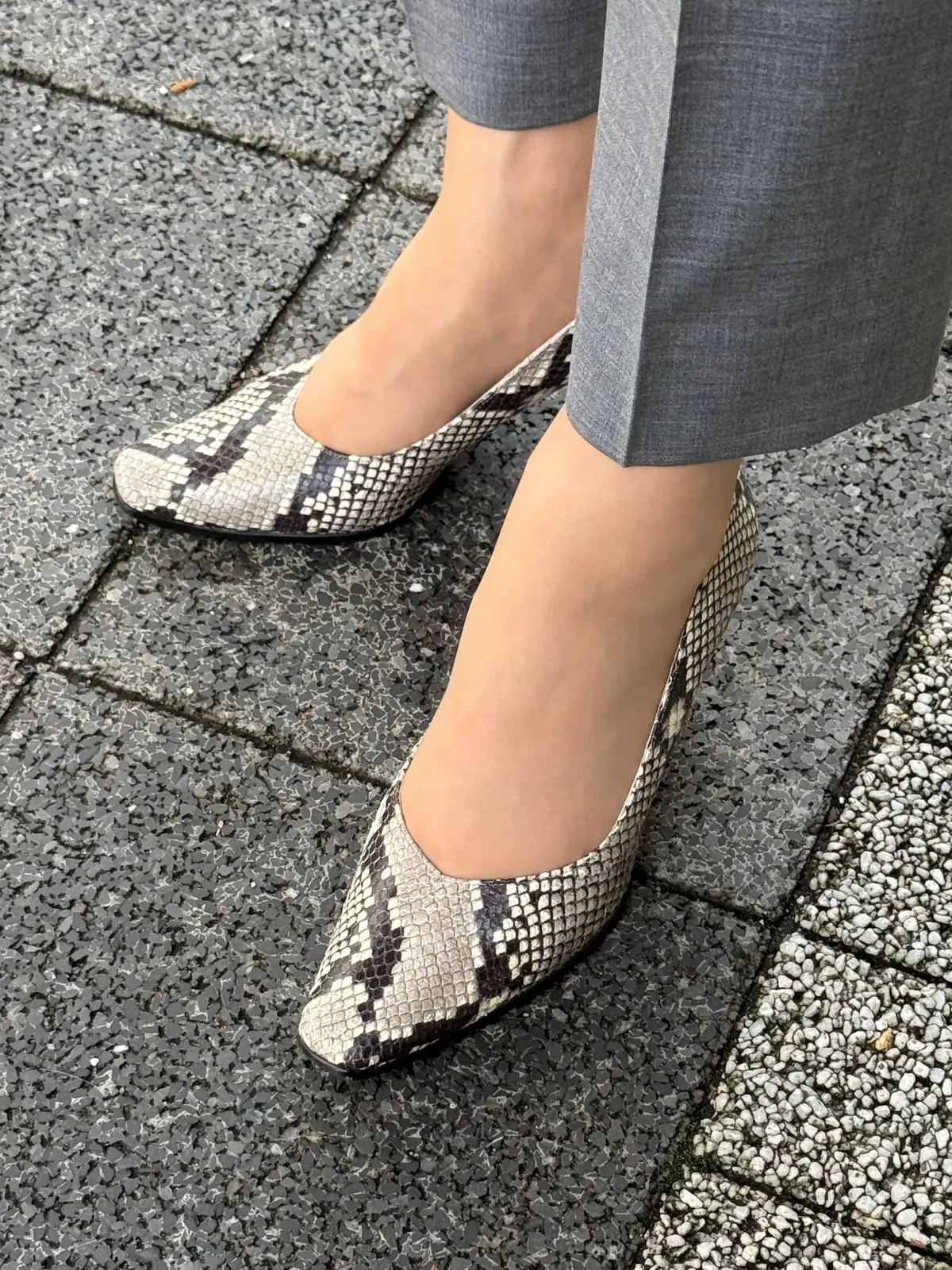 グレーのスラックスを履いた足元。白地に黒と茶色の蛇柄のパンプスを履いている。地面はグレーのタイル模様。