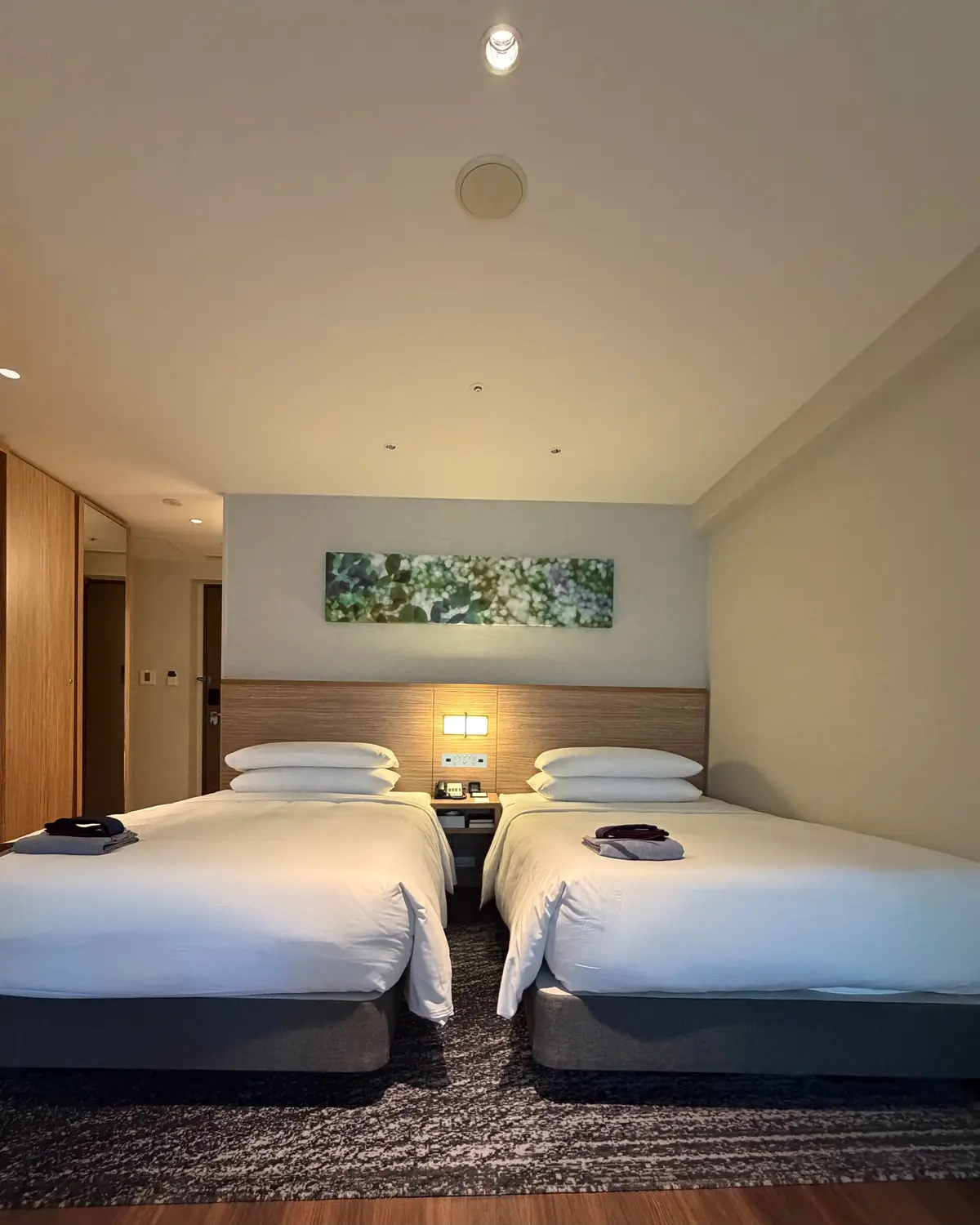 白のシーツをかけたツインベッドが写るホテルの部屋。壁掛けテレビと木製のサイドテーブル、窓際に椅子とテーブルが置かれている。全体的に明るく、シンプルな内装。