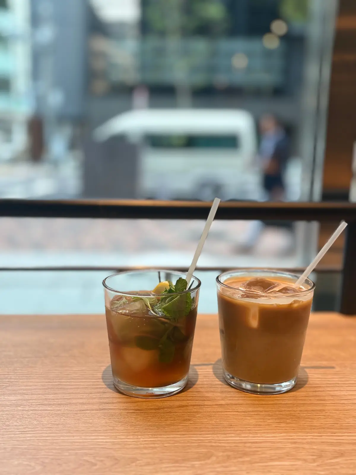 木製のテーブルに置かれた、2つのグラスに入った飲み物。左はミントの葉が飾られた茶色い飲み物、右はミルクを混ぜたような色の飲み物。それぞれにストローが挿さっている。背景には窓から街の風景が見える。