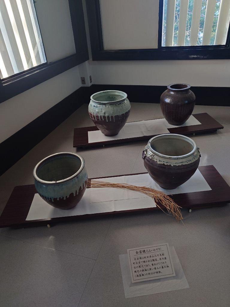 茶室の展示ケース内に置かれた、大小様々な形の陶器が並んでいる。緑青色や茶色がかった釉薬が施された壺や碗が、木製の台に配置されている。背景には窓から庭の緑が見える。