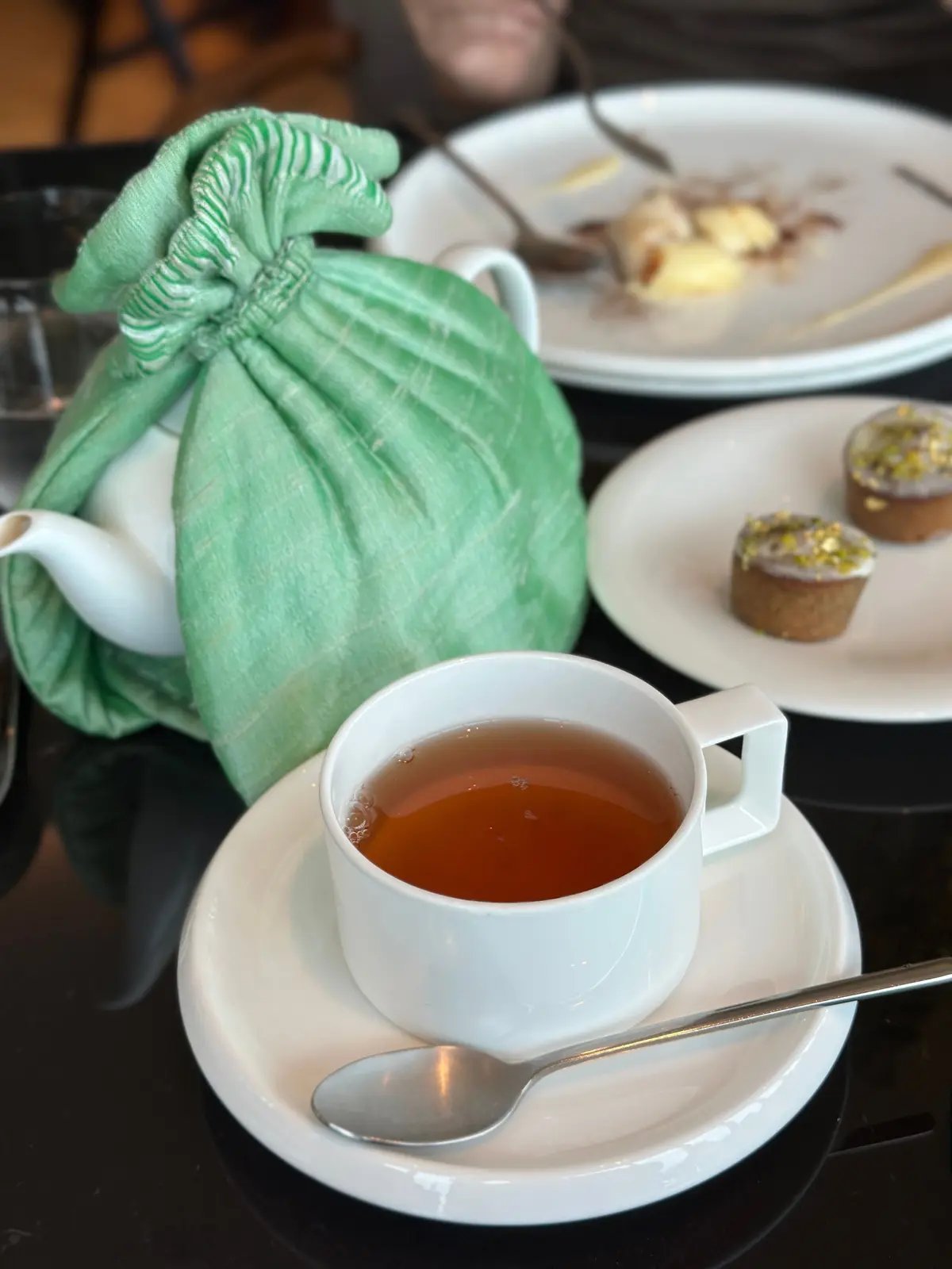 白磁のカップに入った紅茶と、緑色の布で包まれた急須。2つの白い皿には、それぞれ異なる焼き菓子が置かれている。黒いテーブルに置かれたティーセットの様子。