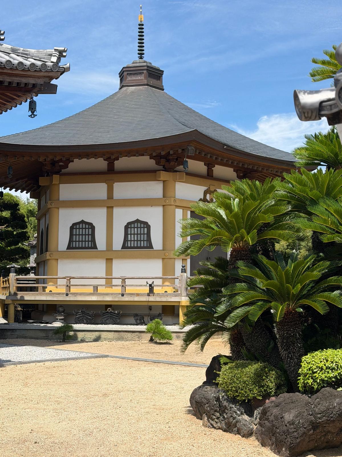 屋根付きの円形の建物。白壁に黄色の縁取り、中央に金色の塔。手前に砂利敷きの庭と緑の植物。青空を背景に、別の建物の屋根の一部が見える。