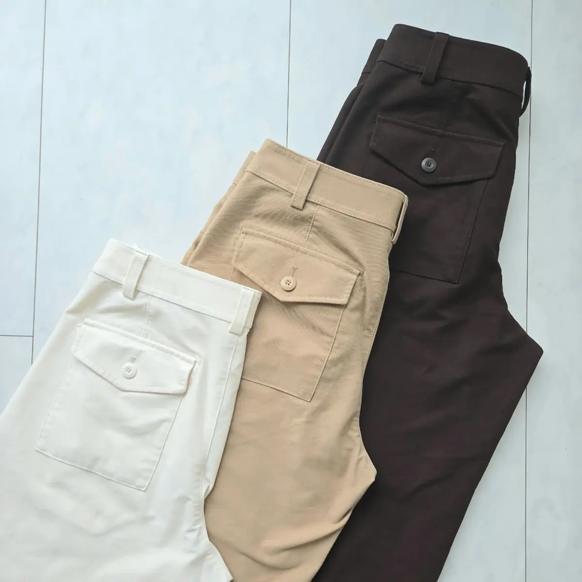 白い、ベージュ、茶色の3本のパンツが畳んで並んでいる。それぞれにフラップ付きのポケットが付いており、ベルトを通すためのループがある。背景は白い床。